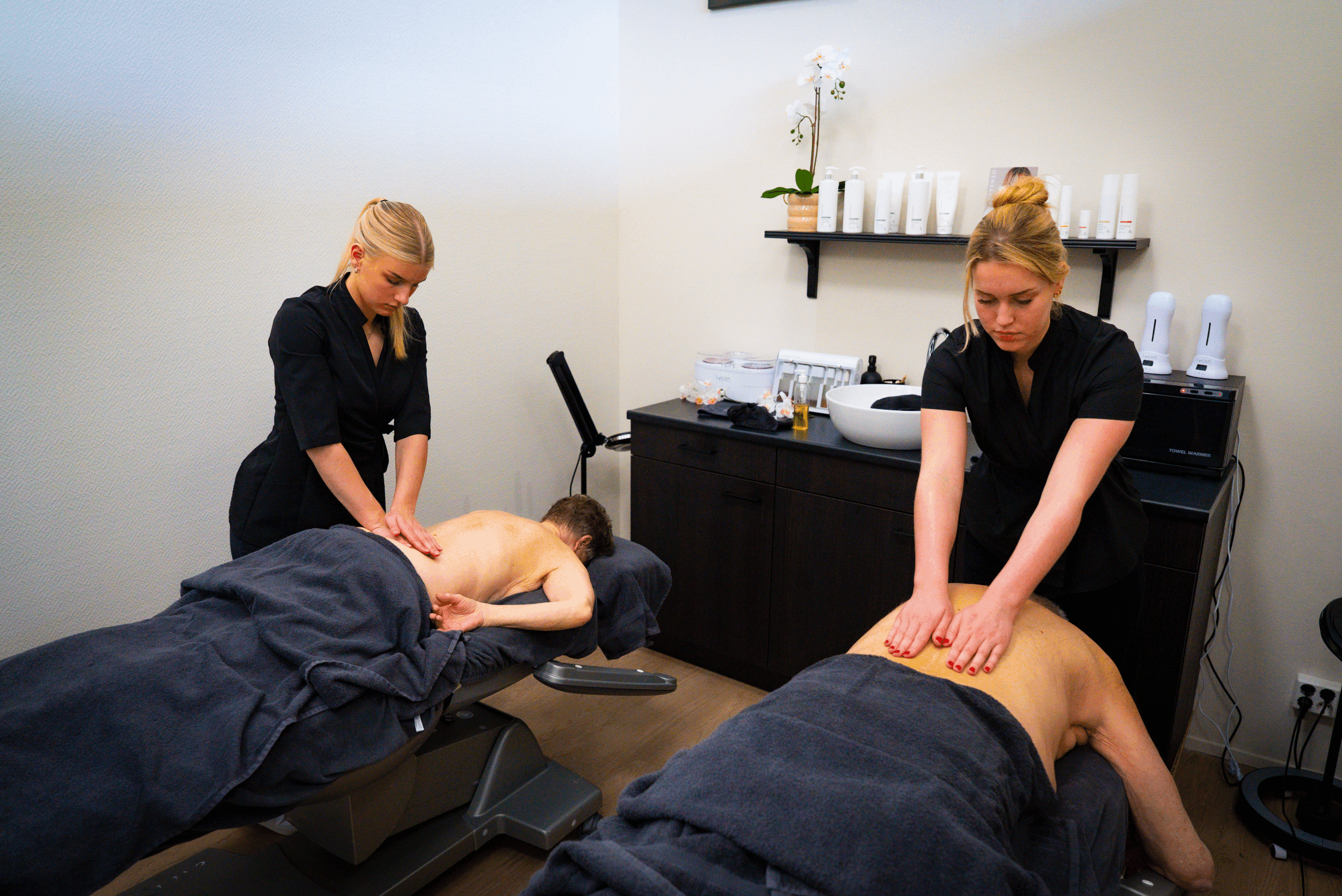 Je ziet een ruimte met 2 mannen op een massagebank die worden gemasseerd door 2 meiden