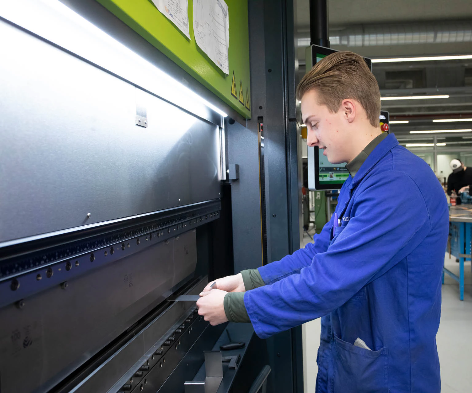 Jongen in een stofjas buigt een staalplaatje in een machine.