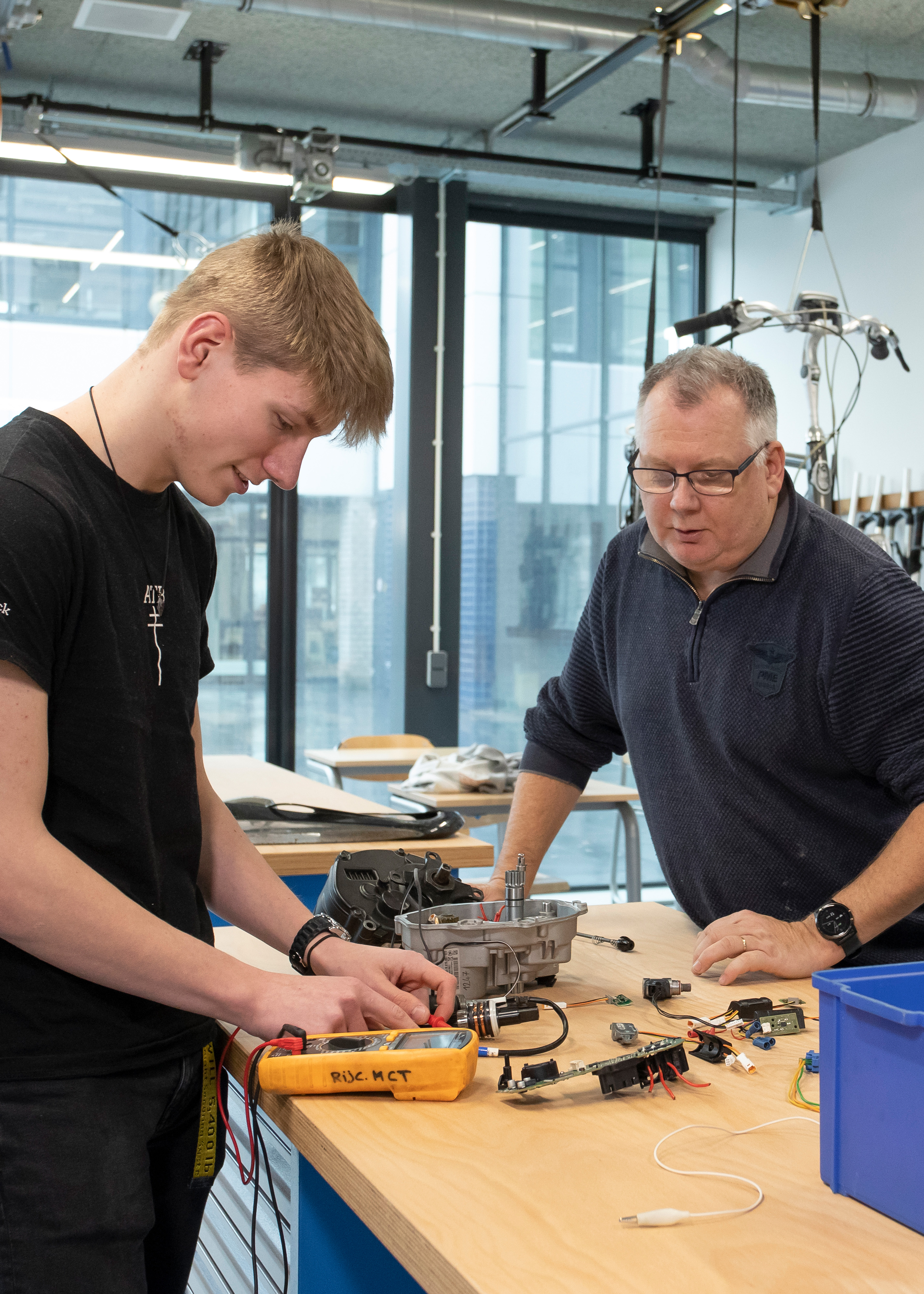 Student is bezig met onderdelen van een fiets op een werkbank terwijl een docent toekijkt