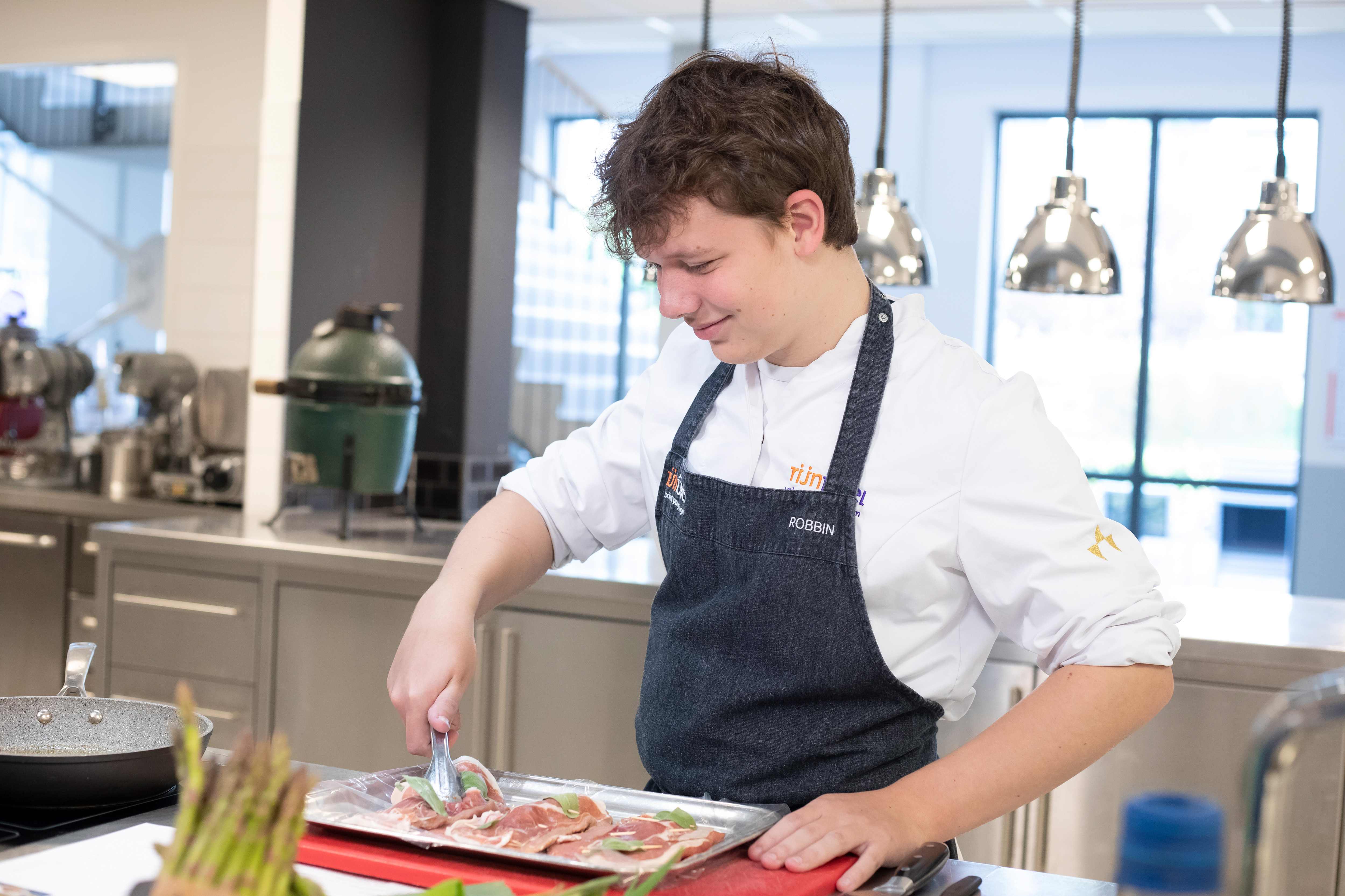 Jongen in kokskleding heeft een tang in de hand en pakt een stuk vlees vast