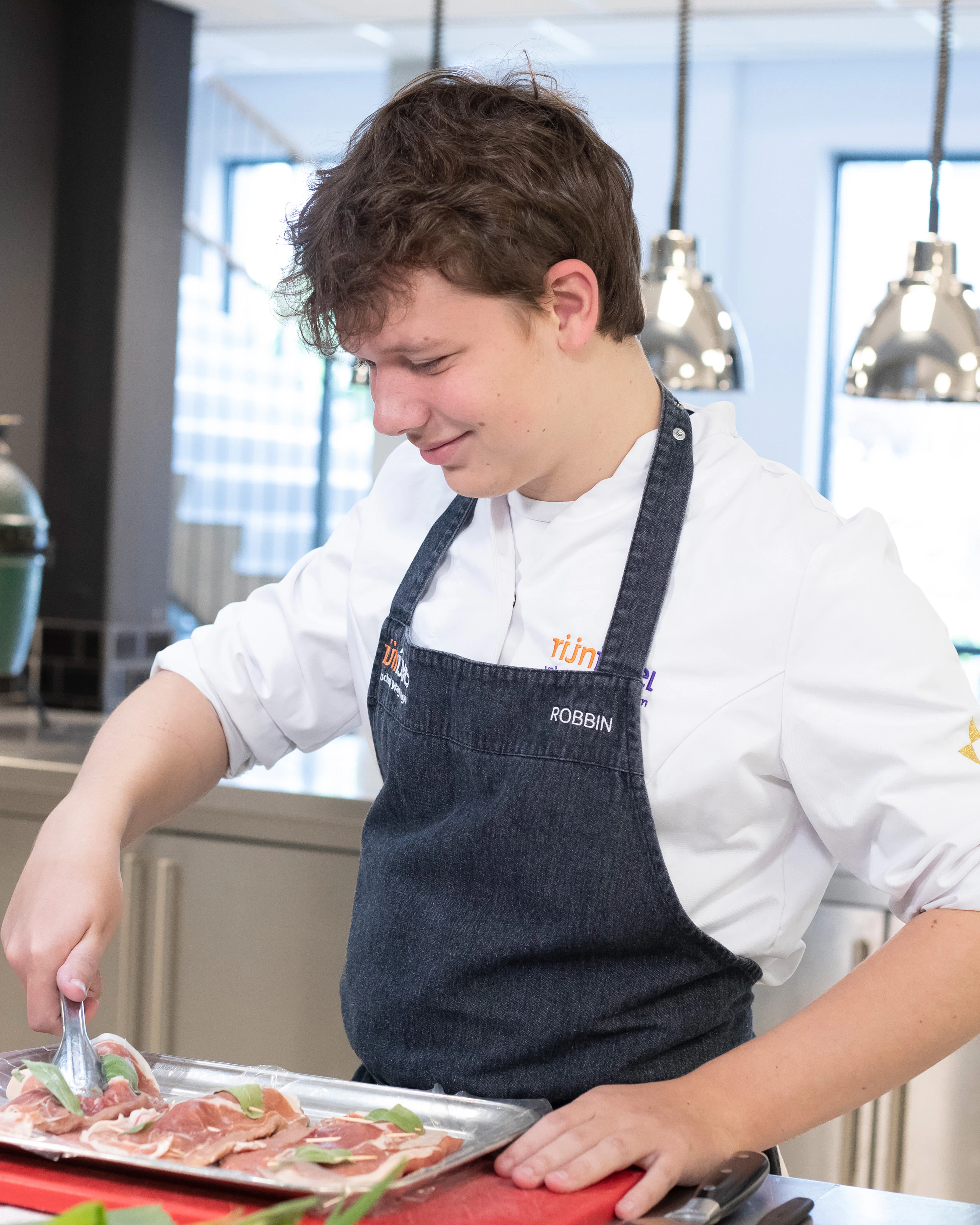 Jongen in kokskleding heeft een tang in de hand en pakt een stuk vlees vast