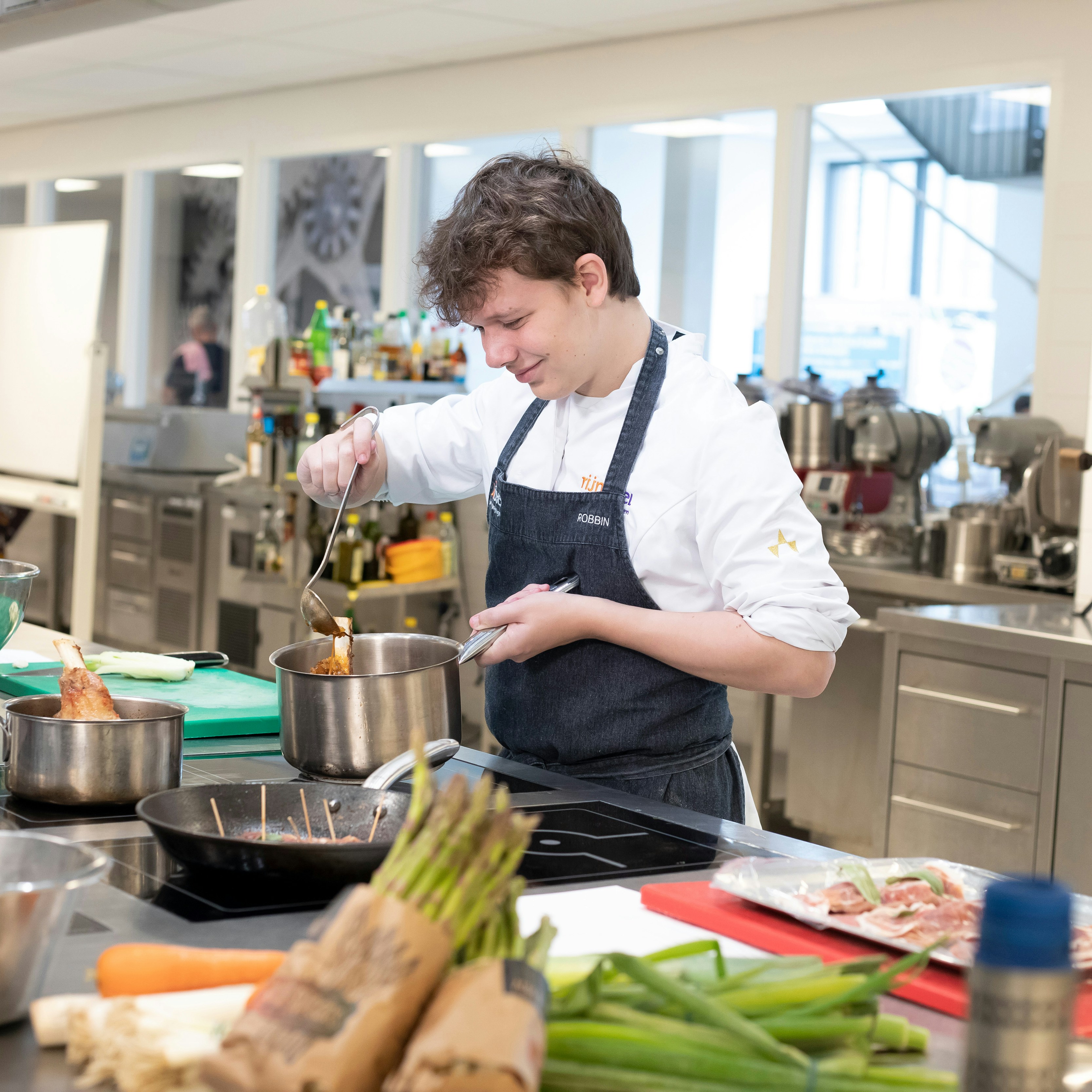 Jongen in kokskleding is aan het koken met een steelpan en heeft een lepel in zijn hand