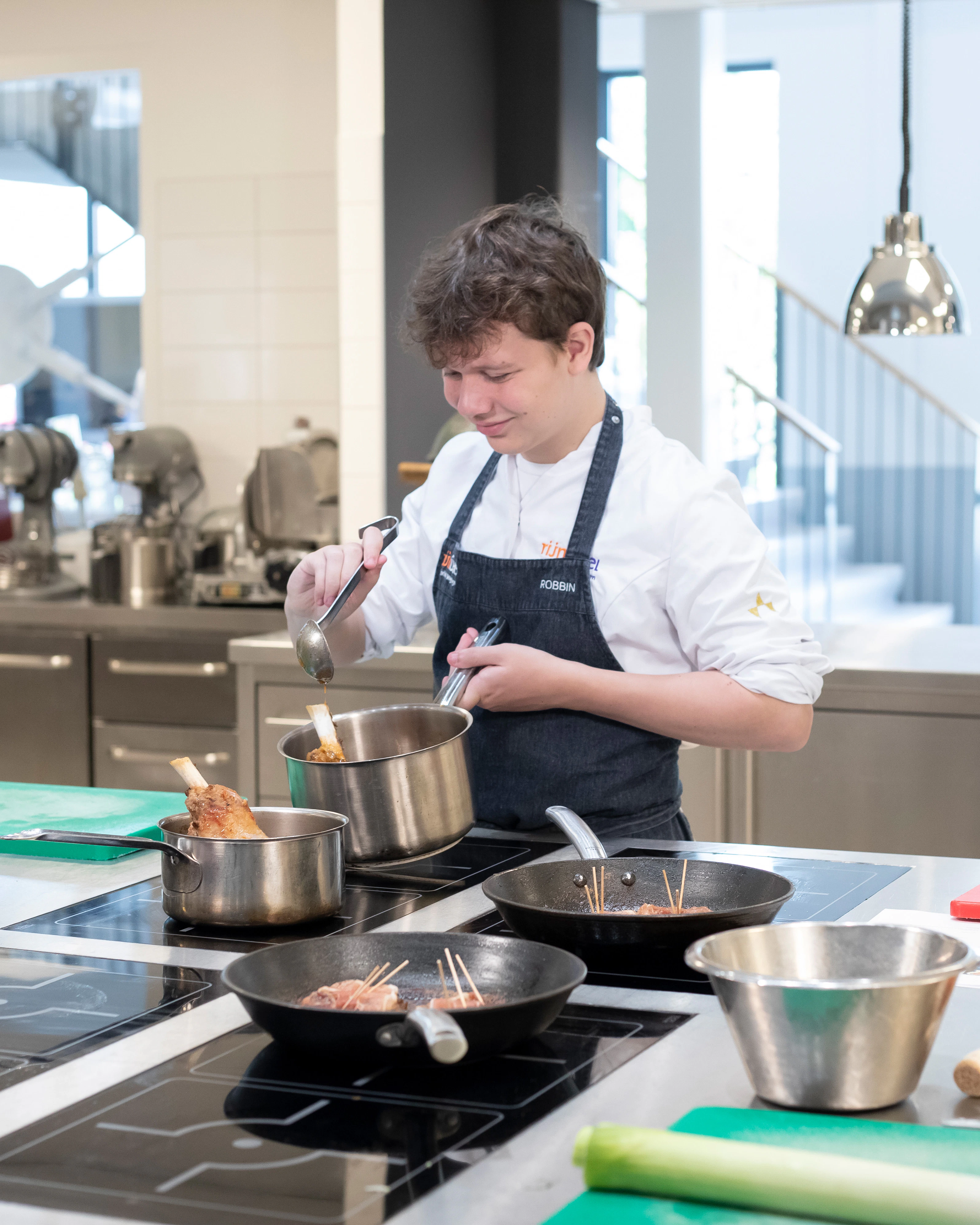 Jongen in kokskleding is aan het koken met een steelpan terwijl hij een lepel vasthoudt