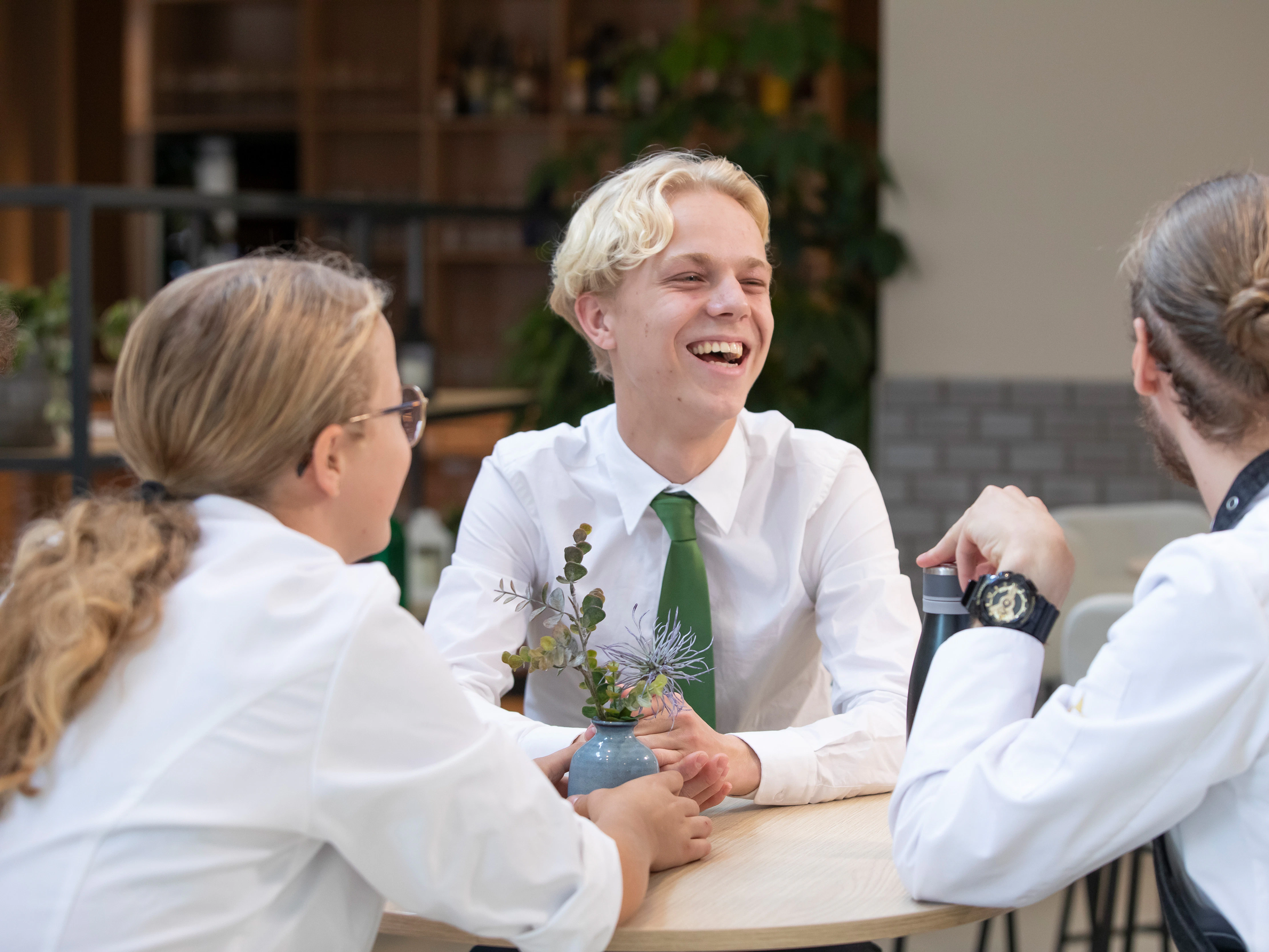 Een meisje en een jongen in kokskleding en een jongen in pak zitten aan tafel in het restaurant. Ze lachen met elkaar.