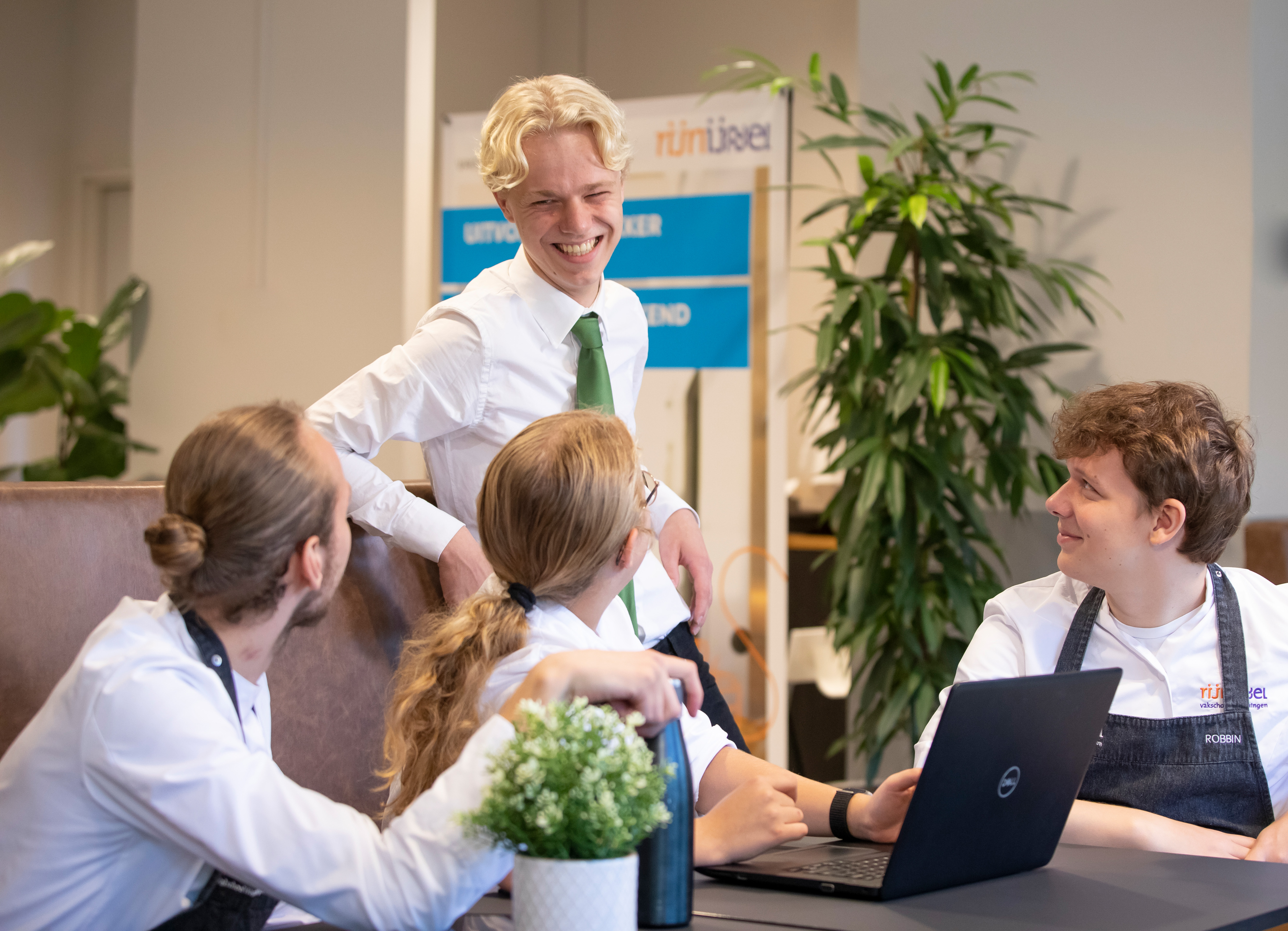 4 studenten in kokskleding kijken elkaar aan. 3 zitten er aan tafel terwijl er 1 staat