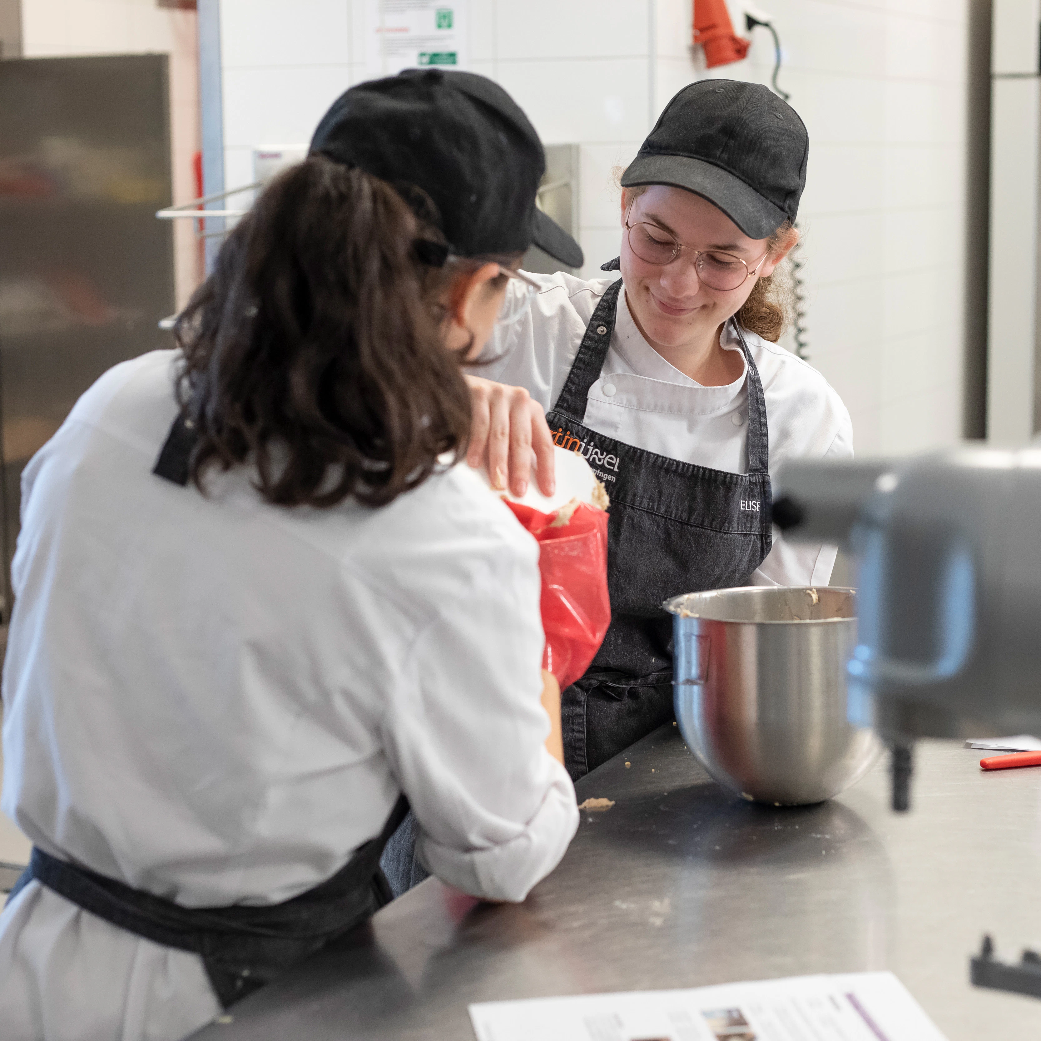 Twee meisjes in bakkerskleding staan bij een mixer en doen beslag in een spuitzak