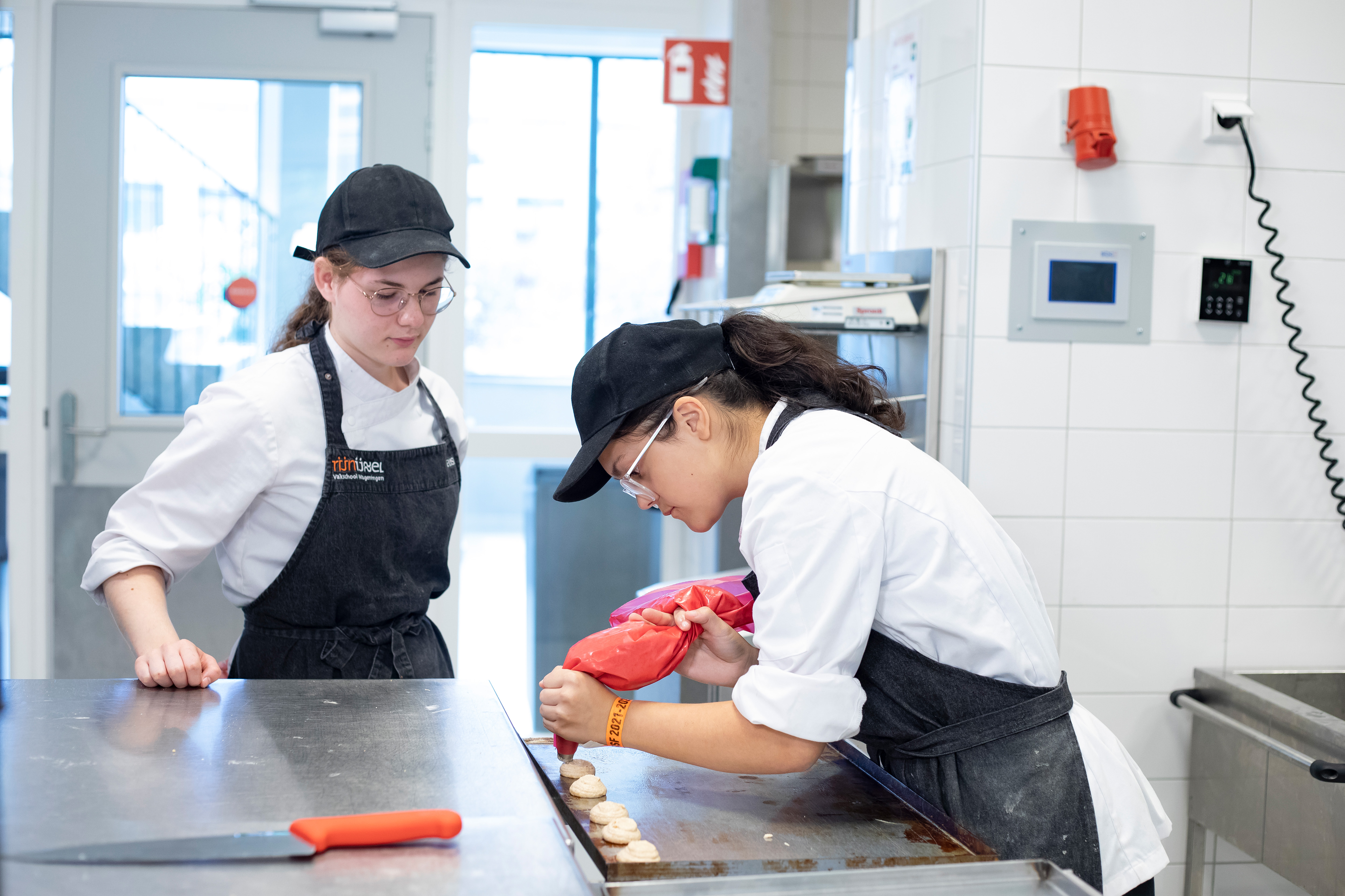 Twee meisjes in bakkerskleding staan in de keuken. Een meisje kijkt toe terwijl het andere meisje met een spuitzak lekkernijen aan het maken is