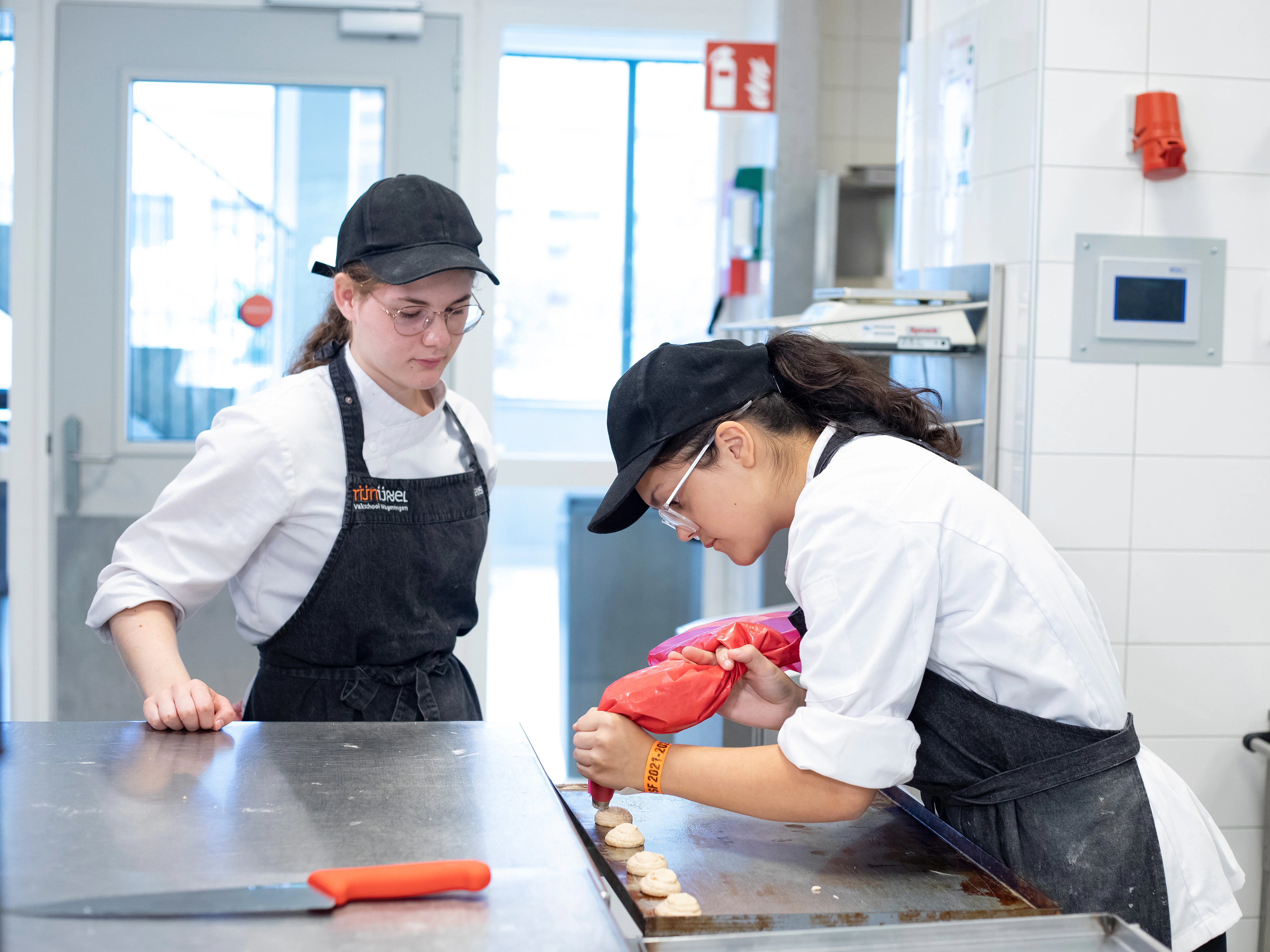 Twee meisjes in bakkerskleding staan in de keuken. Een meisje kijkt toe terwijl het andere meisje met een spuitzak lekkernijen aan het maken is