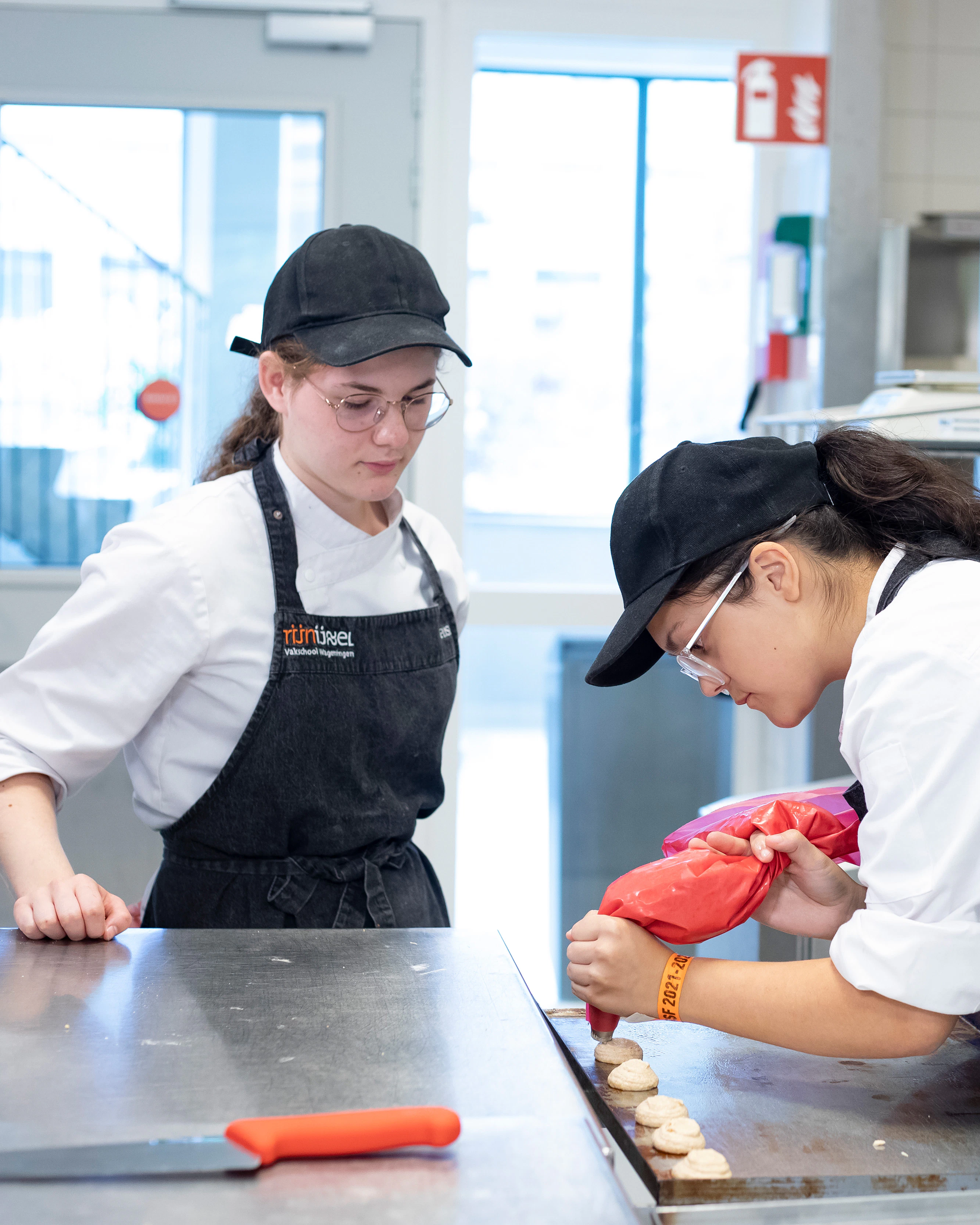 Twee meisjes staan in bakkerskleding in de bakkerij. Een meisje kijkt toe terwijl het andere meisje met de spuitzak lekkernijen aan het maken is