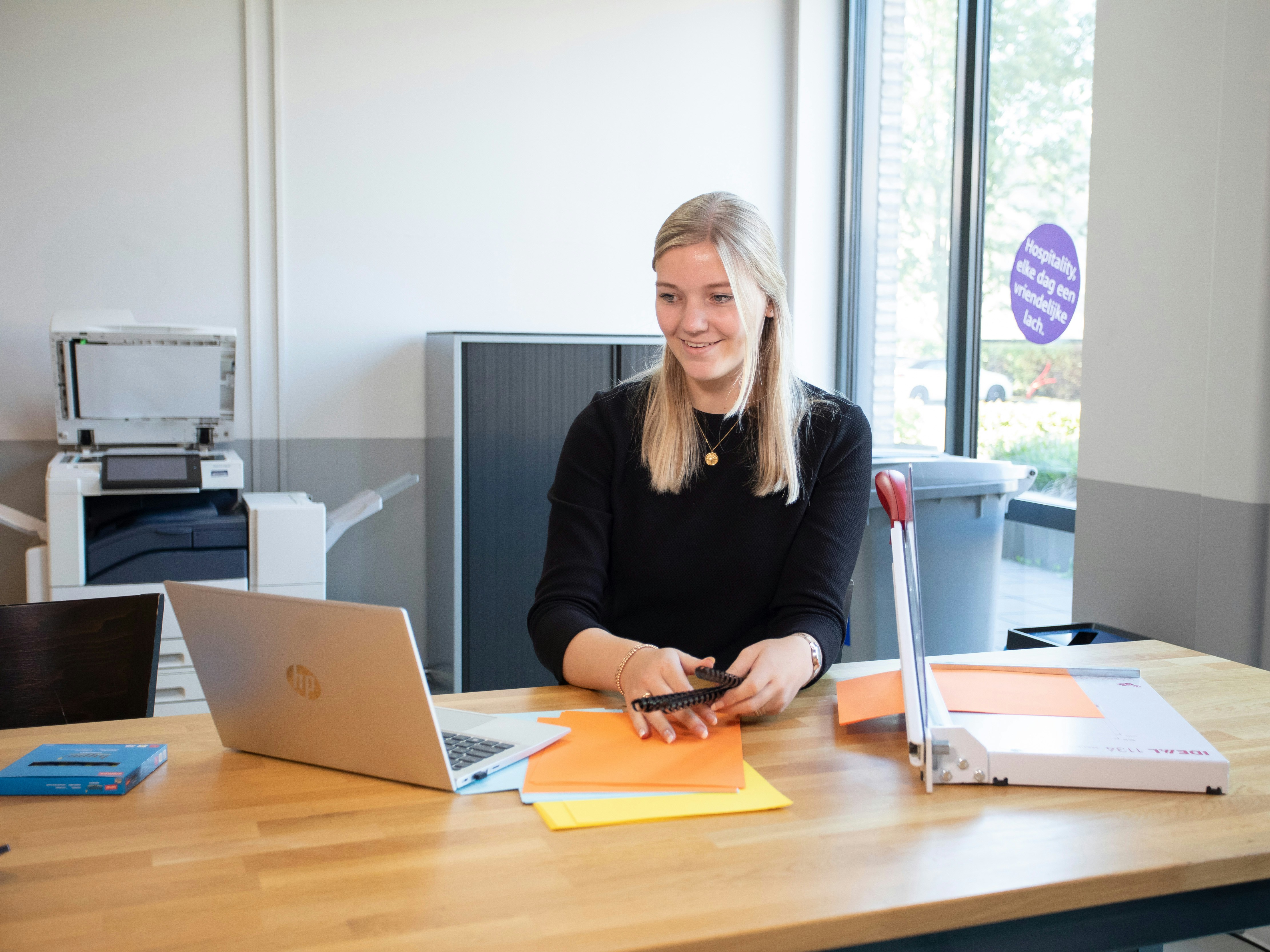 Een meisje zit achter de laptop. Ze kijkt naar de laptop terwijl ze een ringband in haar hand heeft. Op de tafel liggen ook nog gekleurde papieren