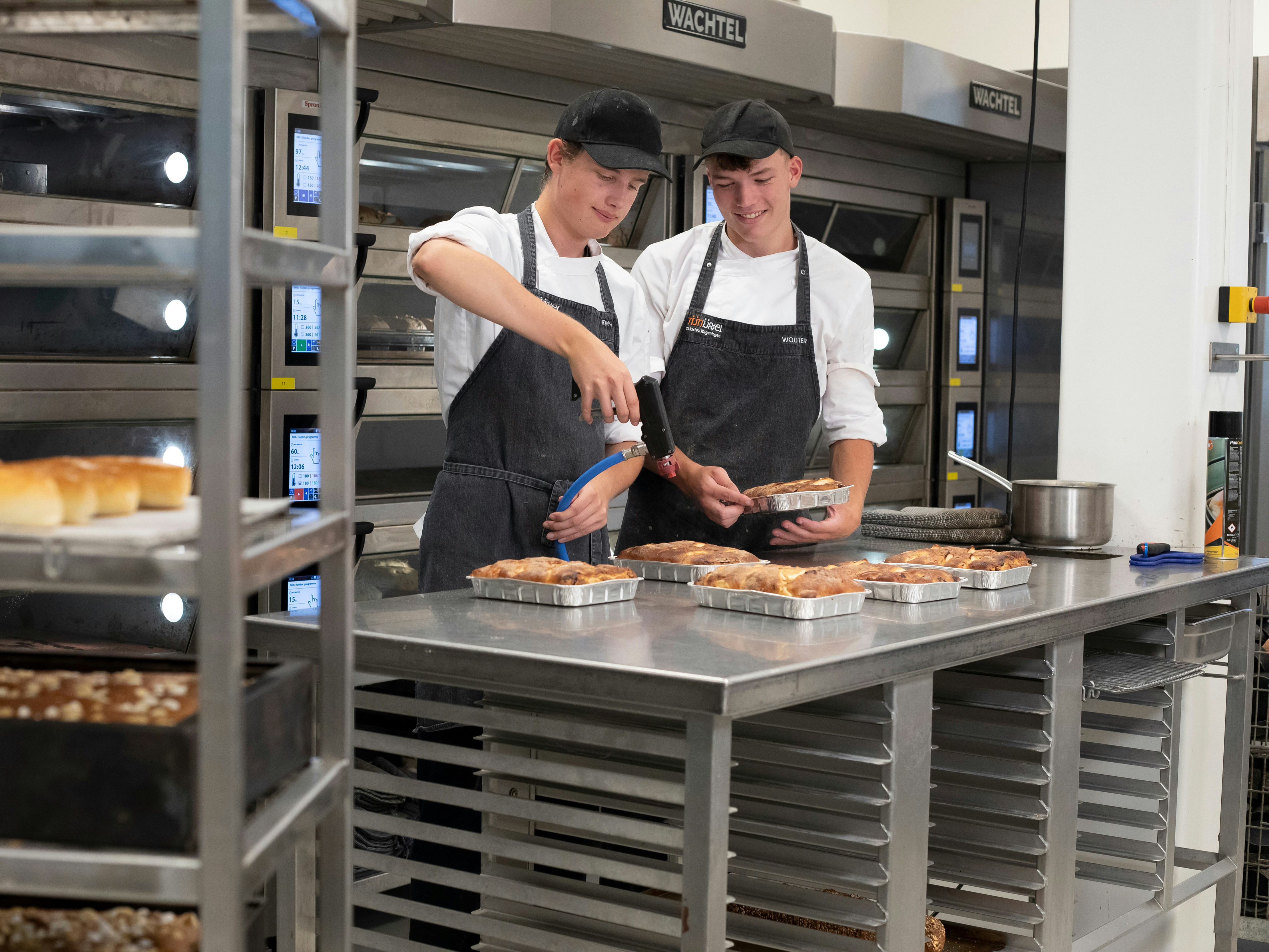 Je ziet twee jongens in de bakkerij met bakkerskleding aan. Op de voorgrond zie je een rek met broden en op de achtergrond zie je de jongens broodgerechten bewerken met een brander