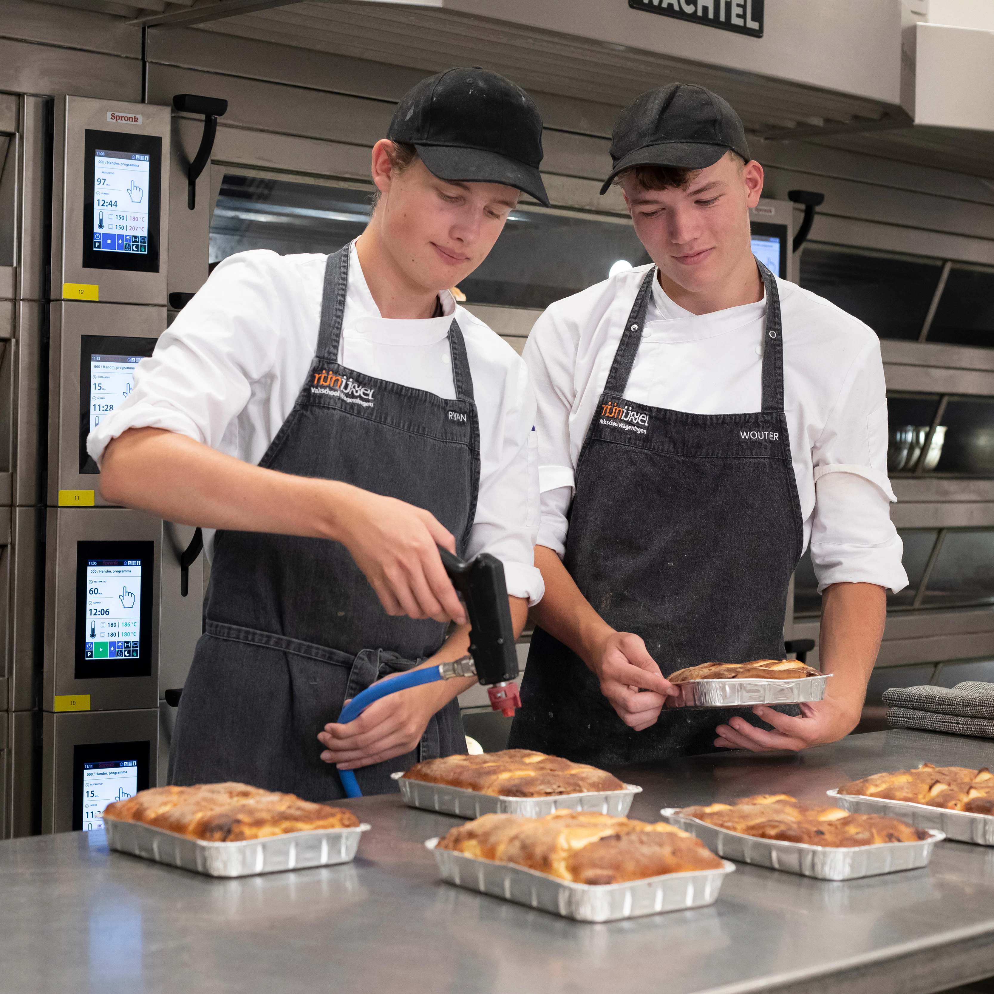 Twee jongens in bakkerskleding staan in de bakkerij voor de oven. Een jongen kijkt toe terwijl de andere jongen 6 broodgerechten aan het bewerken is met een brander
