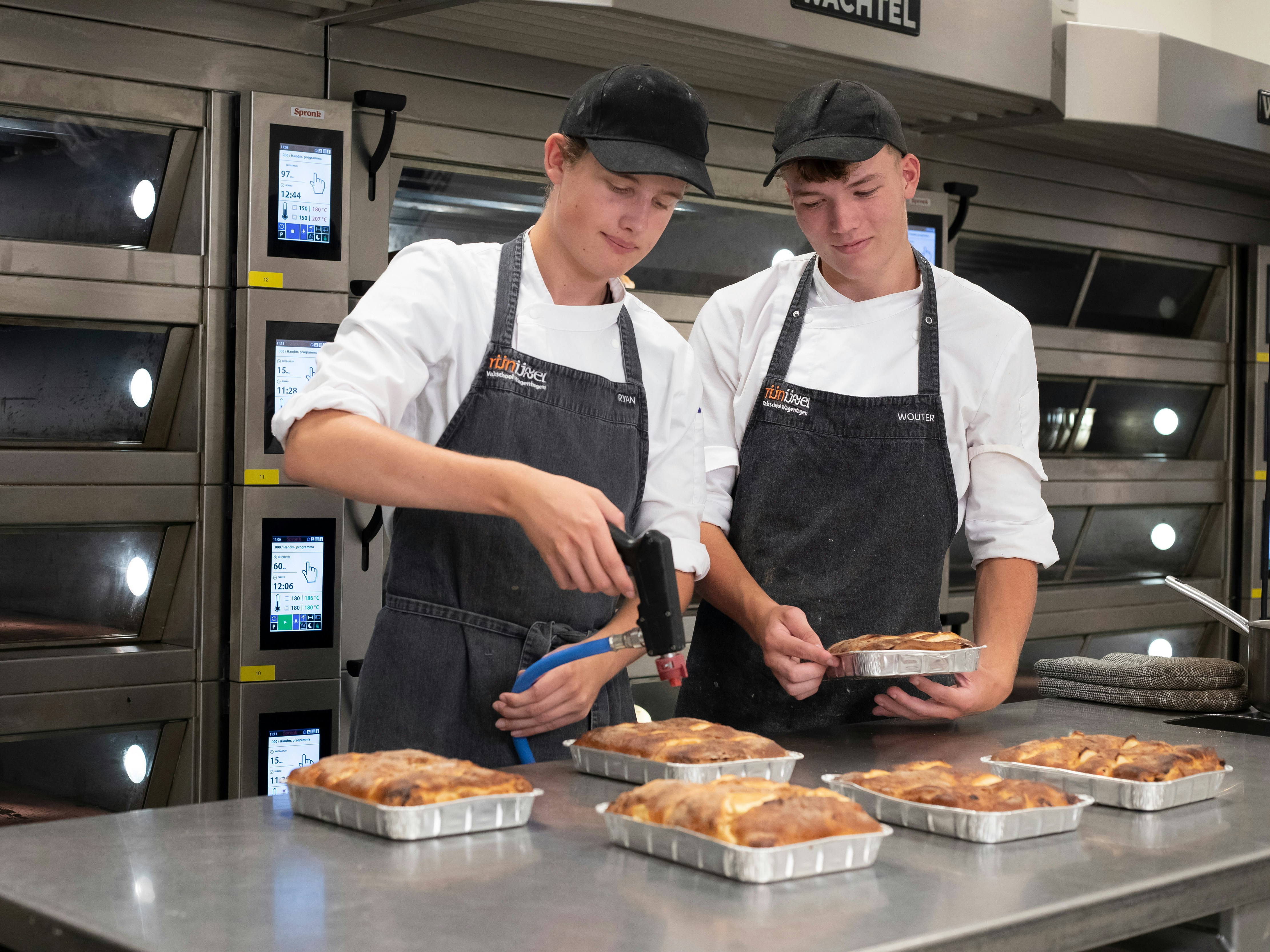 Twee jongens in bakkerskleding staan in de bakkerij voor de oven. Een jongen kijkt toe terwijl de andere jongen 6 broodgerechten aan het bewerken is met een brander