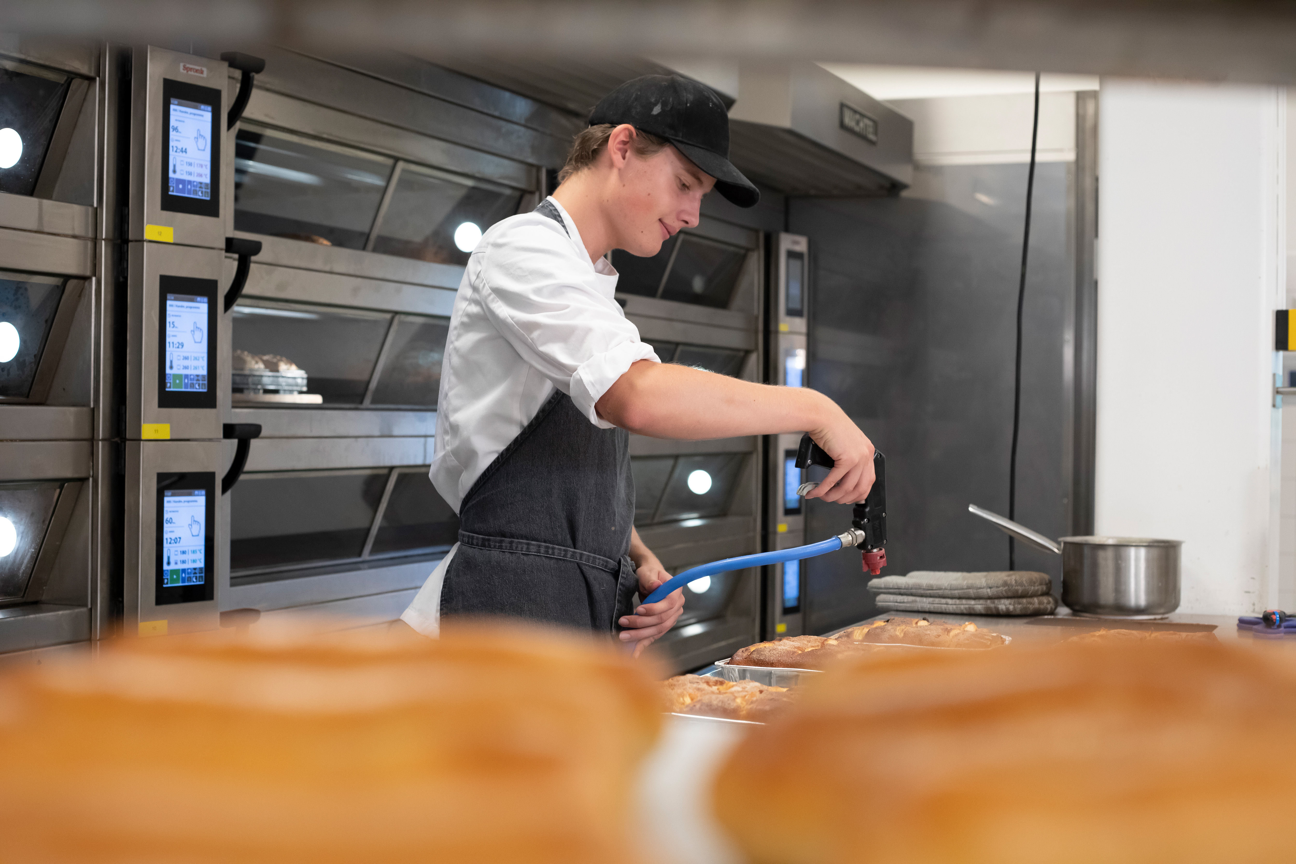 Op de voorgrond zie je een rek met broden en op de achtergrond zie je een jongen in bakkerskleding met een brander broodgerechten bewerken