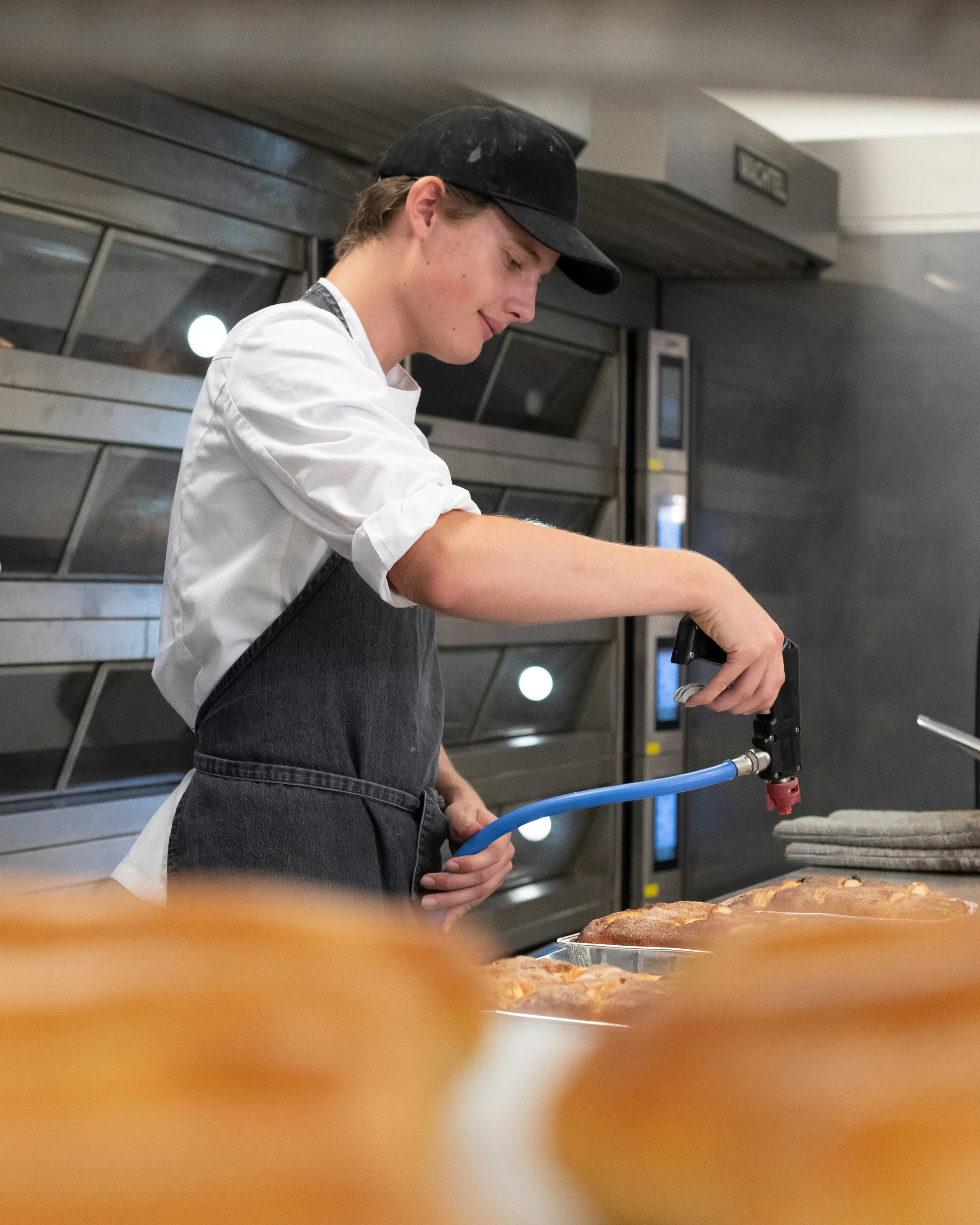 Op de voorgrond zie je een rek met broden en op de achtergrond zie je een jongen in bakkerskleding met een brander broodgerechten bewerken