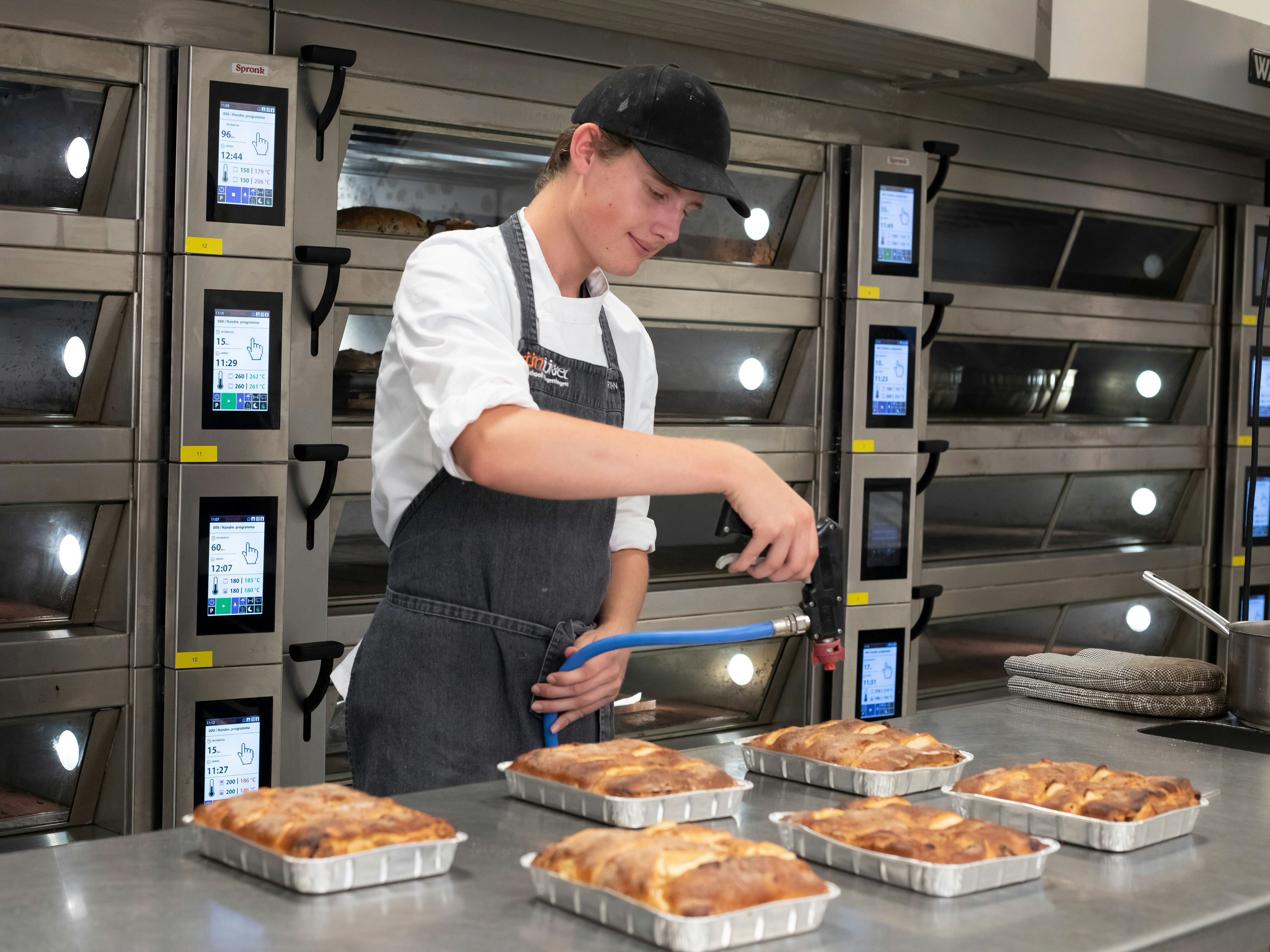 Jongen staat in bakkerskleding in de bakkerij met zes broodgerechten voor hem die hij aan het bewerken is met een brander
