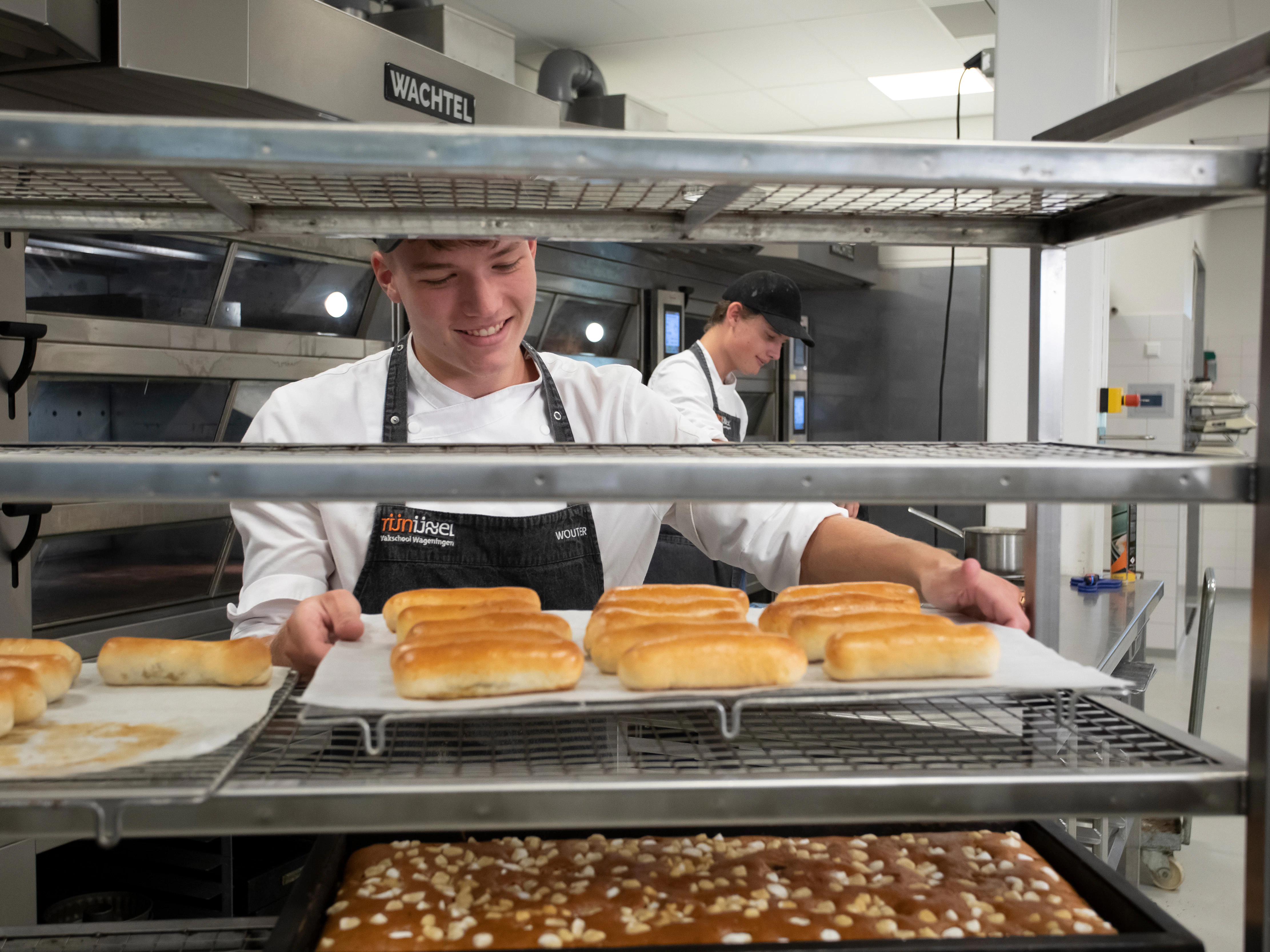 Jongen in bakkerskleding in een bakkerij staat achter een rek waar broden en cakejes liggen