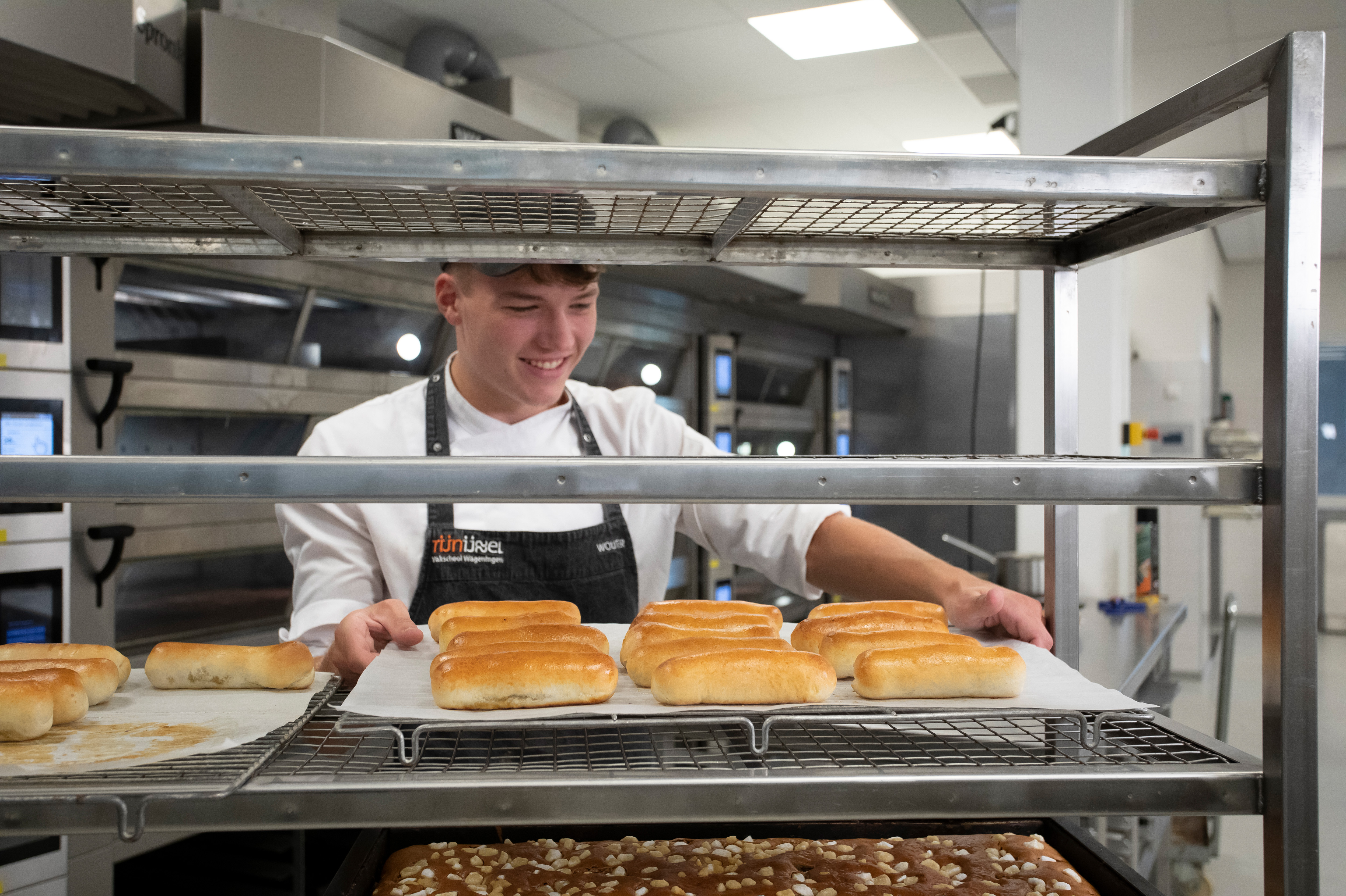 Je ziet een rek met broden en cakes. Daar staat een jongen in bakkerskleding achter die de broden uit het rek haalt