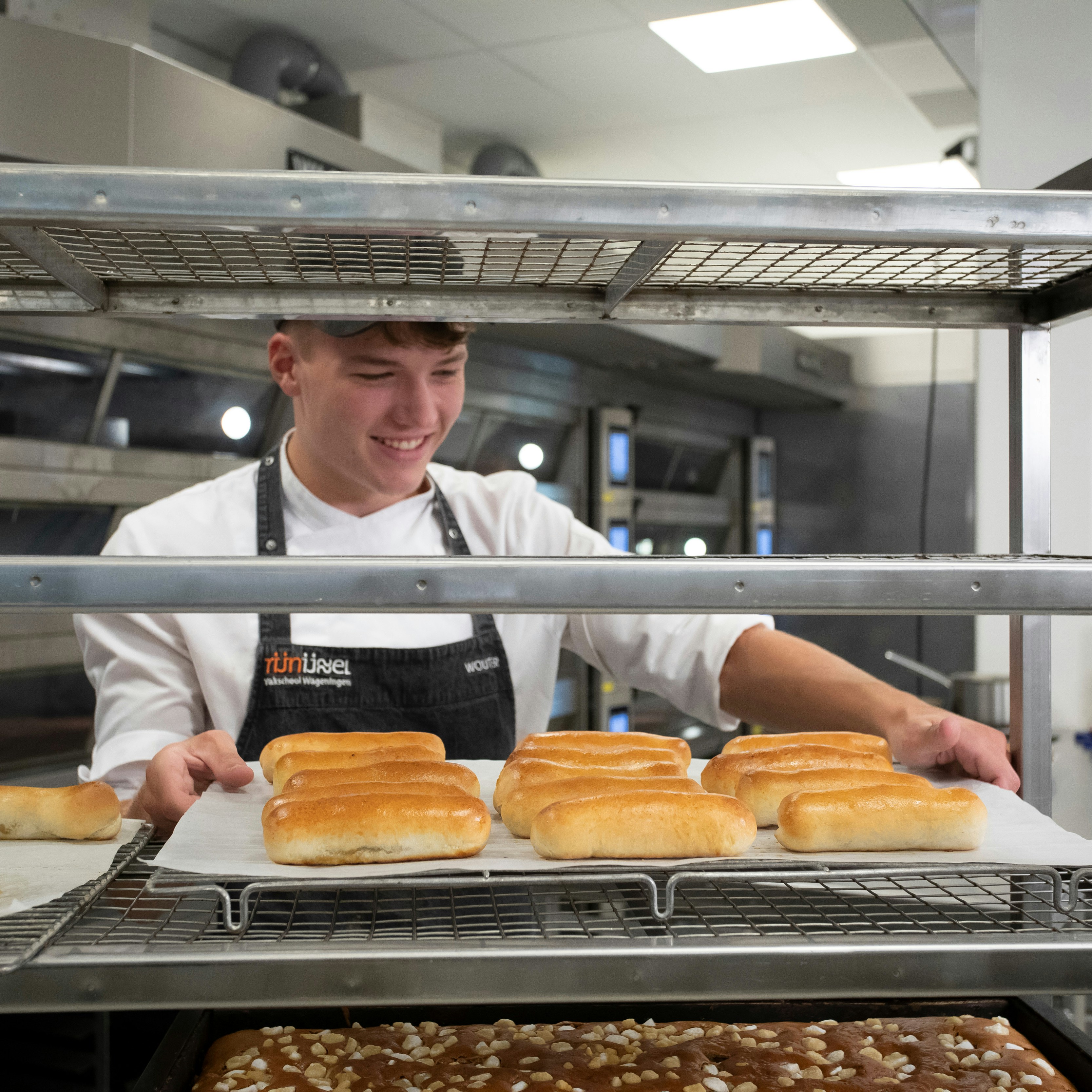 Jongen in bakkerskleding in een bakkerij staat achter een rek waar broden en cakejes liggen