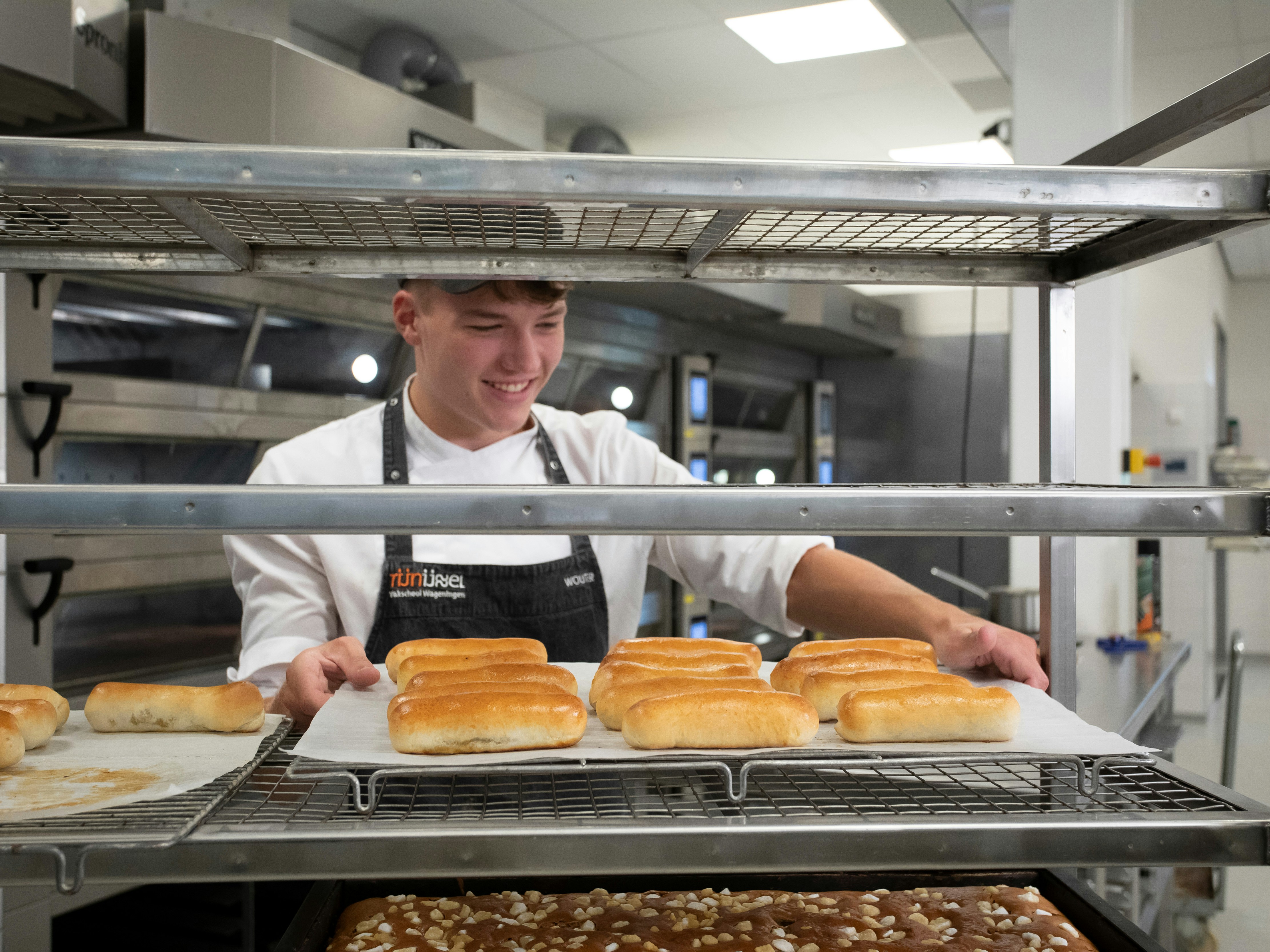 Jongen in bakkerskleding in een bakkerij staat achter een rek waar broden en cakejes liggen