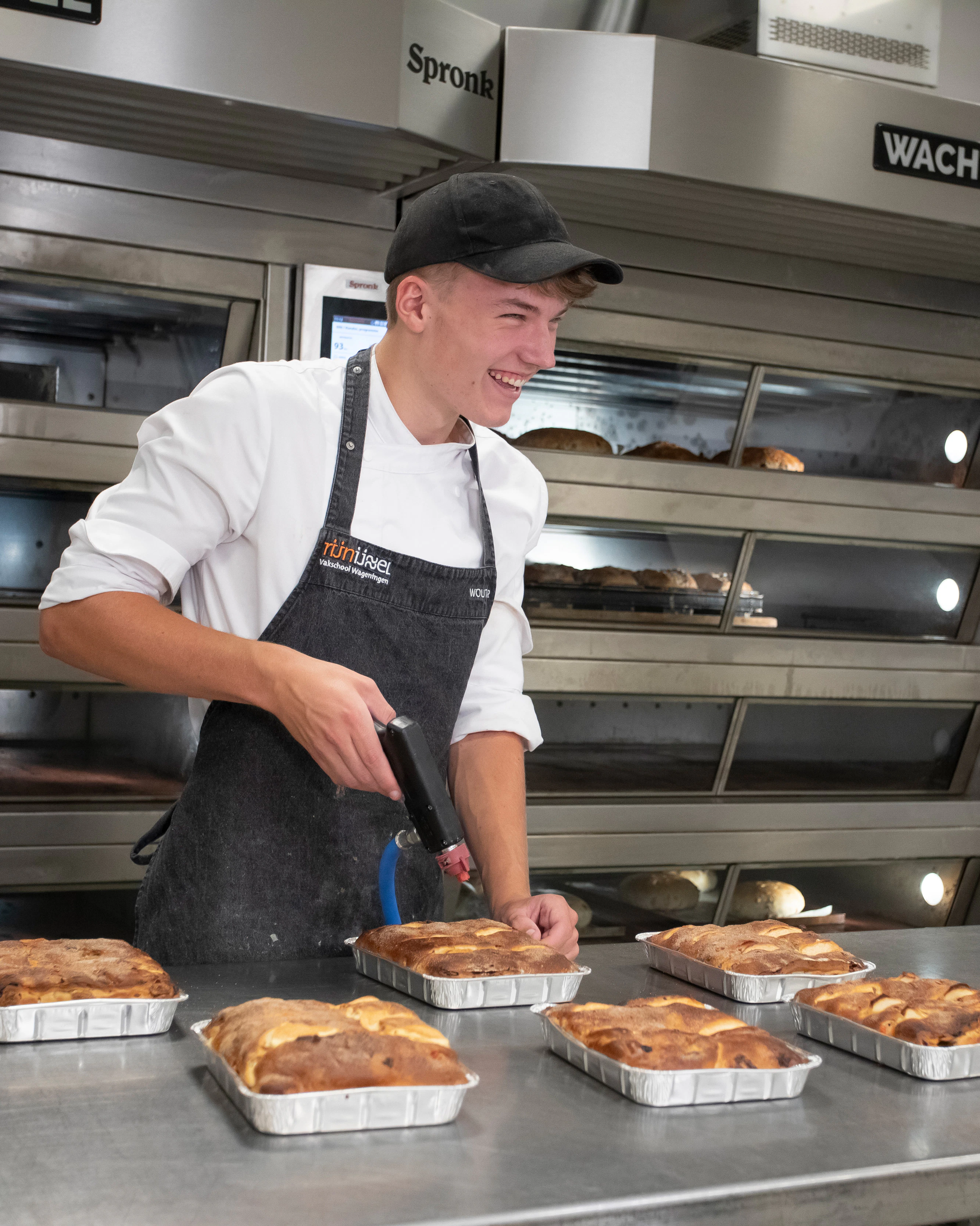 Je ziet een jongen in bakkerskleding in de bakkerij met 6 broodgerechten voor zich. Die is hij aan het bewerken met een brander
