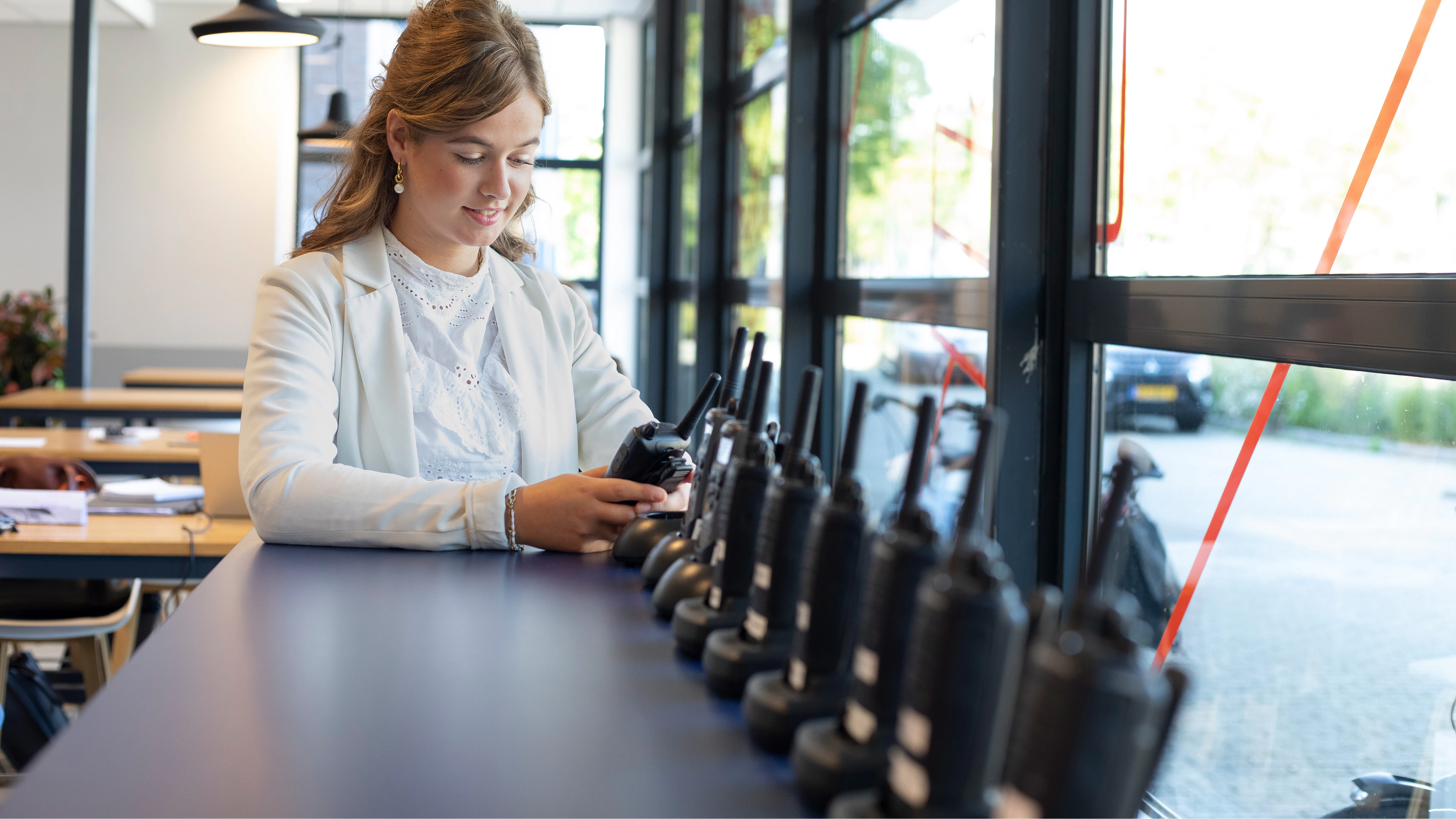 รรฉn meisje in een wit pak. staat bij een kast waarop walkie talkie's staan. Ze heeft er รฉรฉn vast en kijkt daarnaar.
