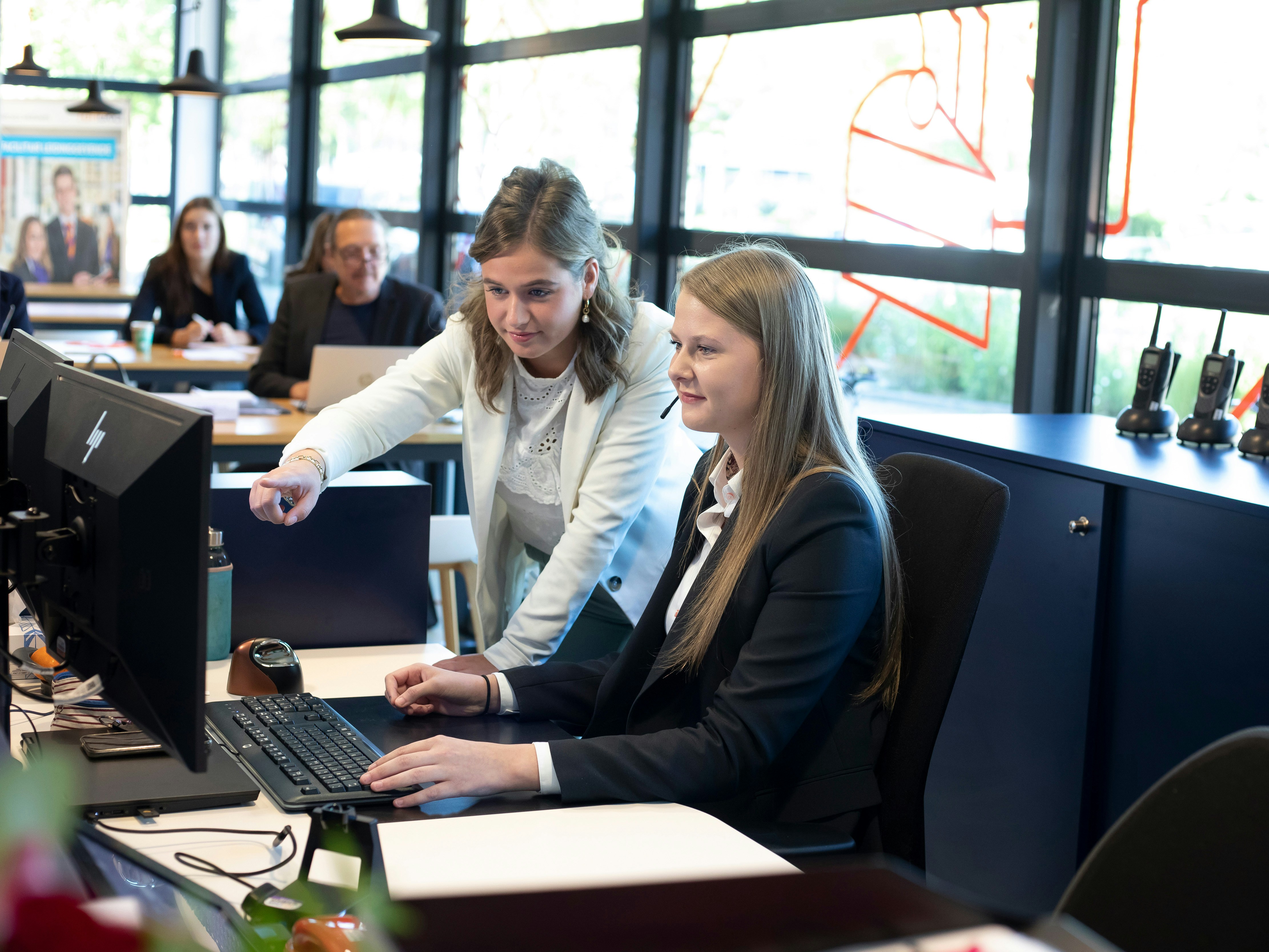 Een meisje in een net pak zit achter de computer terwijl er naast een een ander meisje naar het beeldscherm wijst en haar iets laat zien