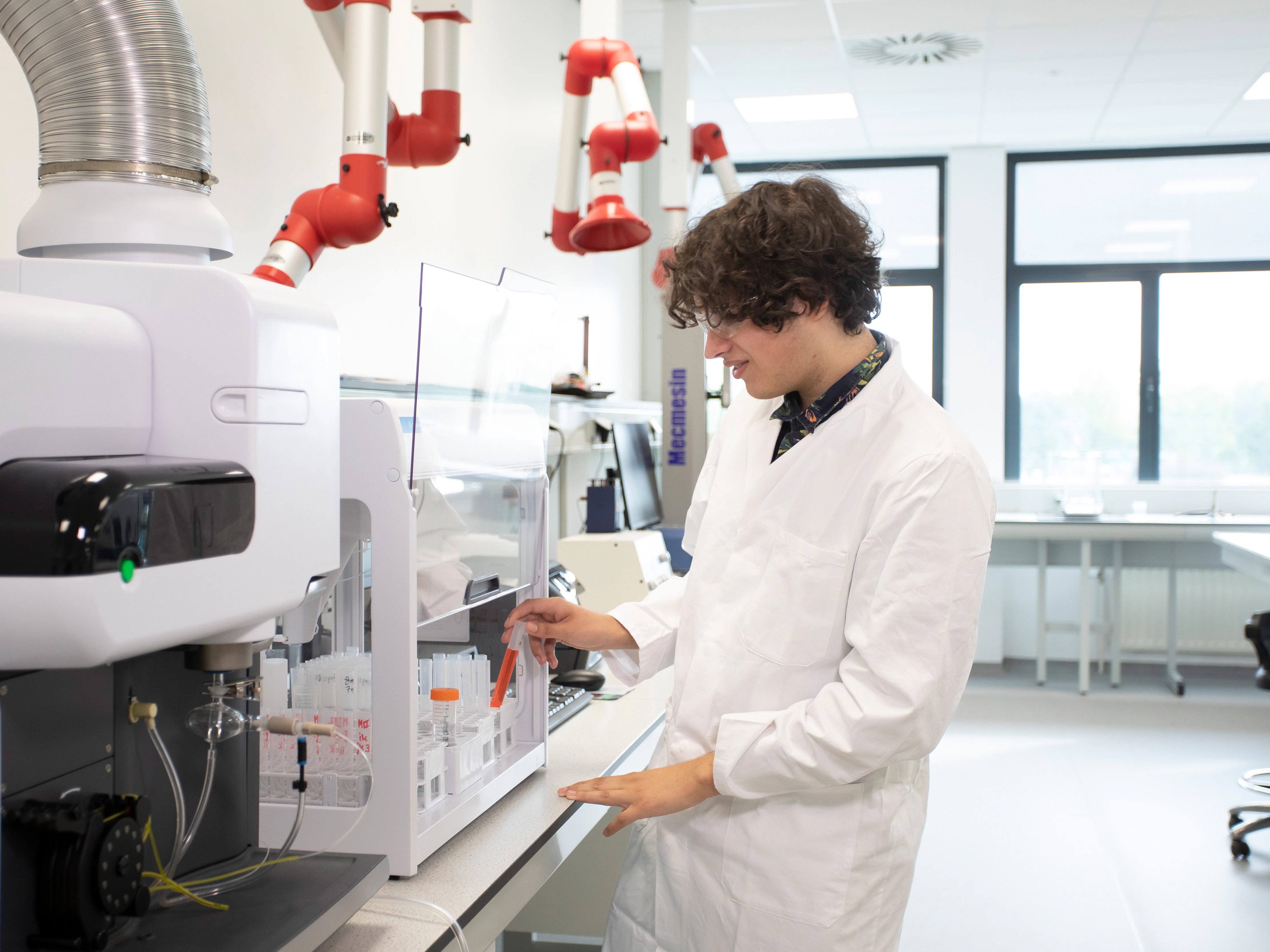 Jongen in een witte laboratoriumjas is bezig bij een machine en heeft oranje chemische vloeistoffen in buisjes