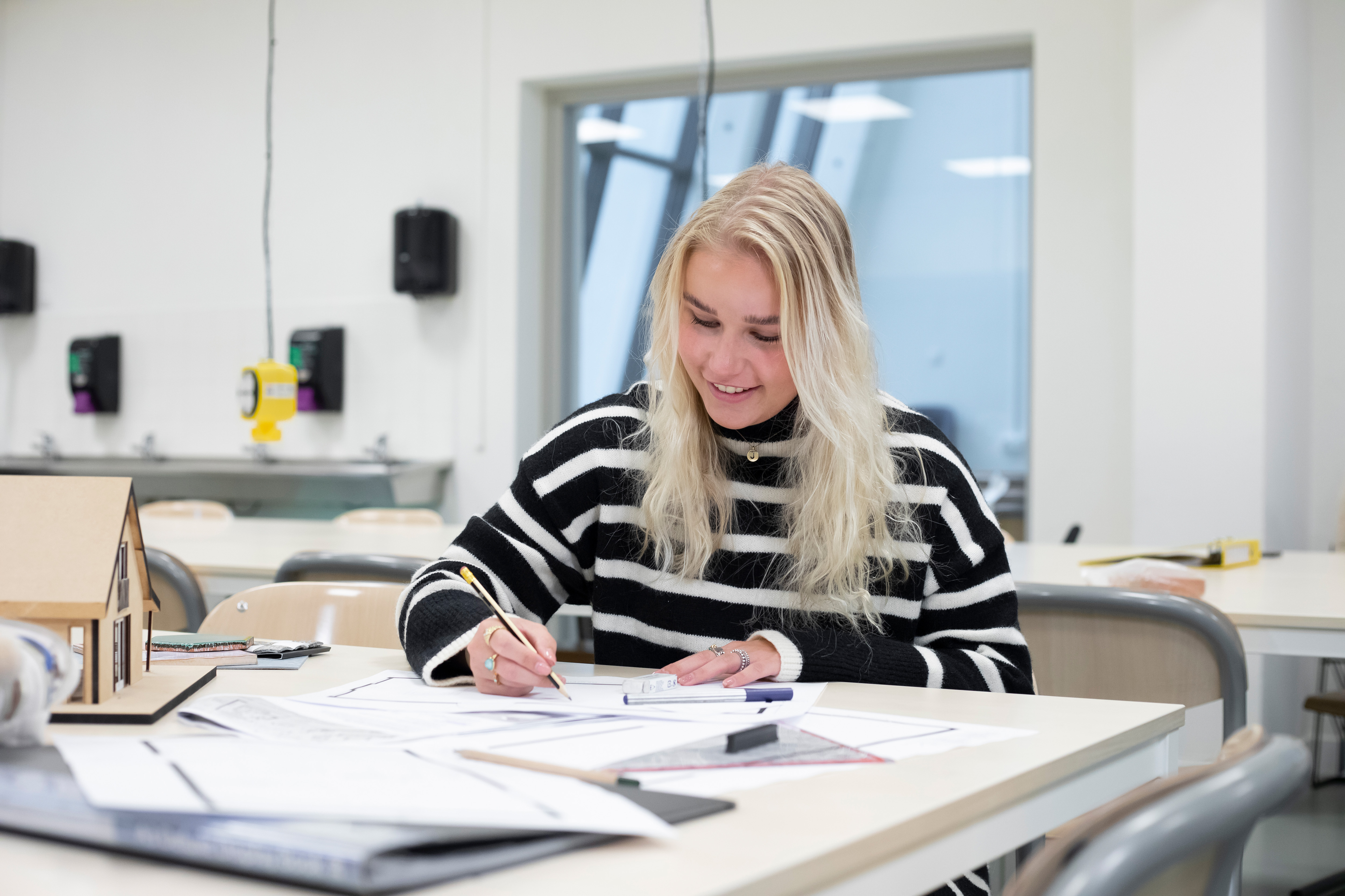 Meisje zit aan een tafel en is aan het tekenen. Op de tafel ligt voor de rest nog papier en er staat een maquette