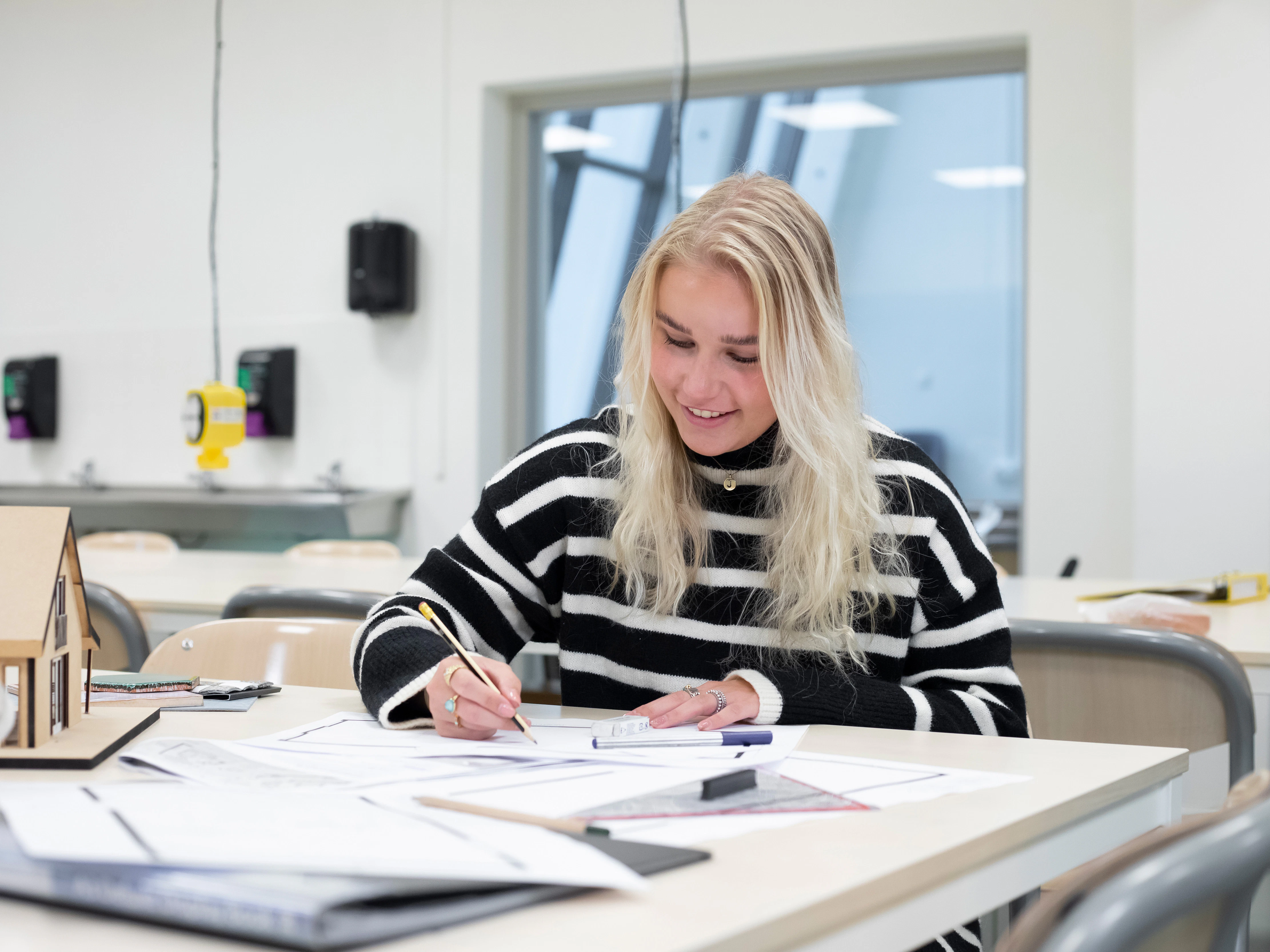 Meisje zit aan een tafel en is aan het tekenen. Op de tafel ligt voor de rest nog papier en er staat een maquette