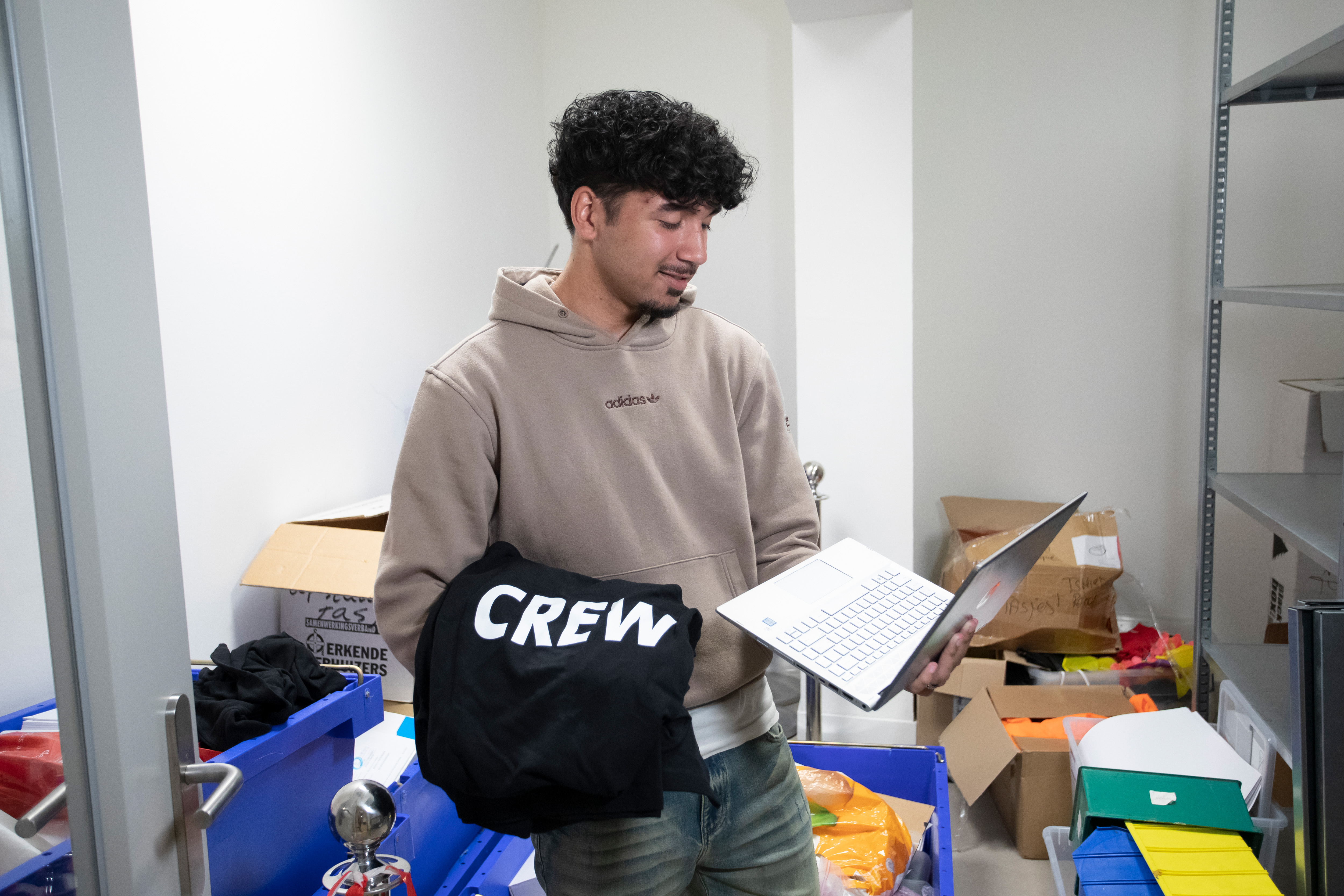 Jongen met een bruine trui staat in een hok met veel spullen. In zijn hand heeft hij een laptop en over zijn andere arm hangt een trui met het woord crew erop