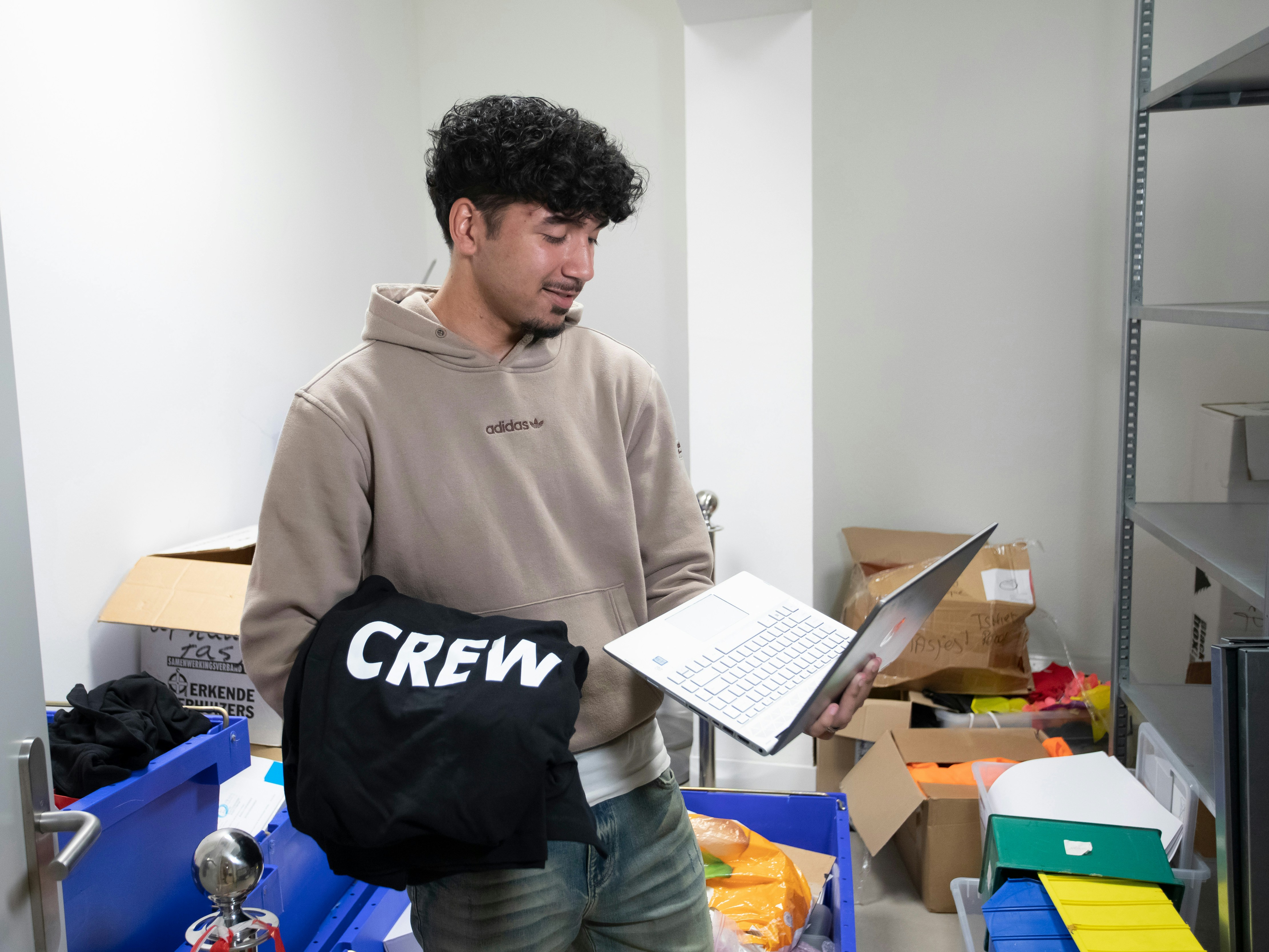 Jongen met een bruine trui staat in een hok met veel spullen. In zijn hand heeft hij een laptop en over zijn andere arm hangt een trui met het woord crew erop
