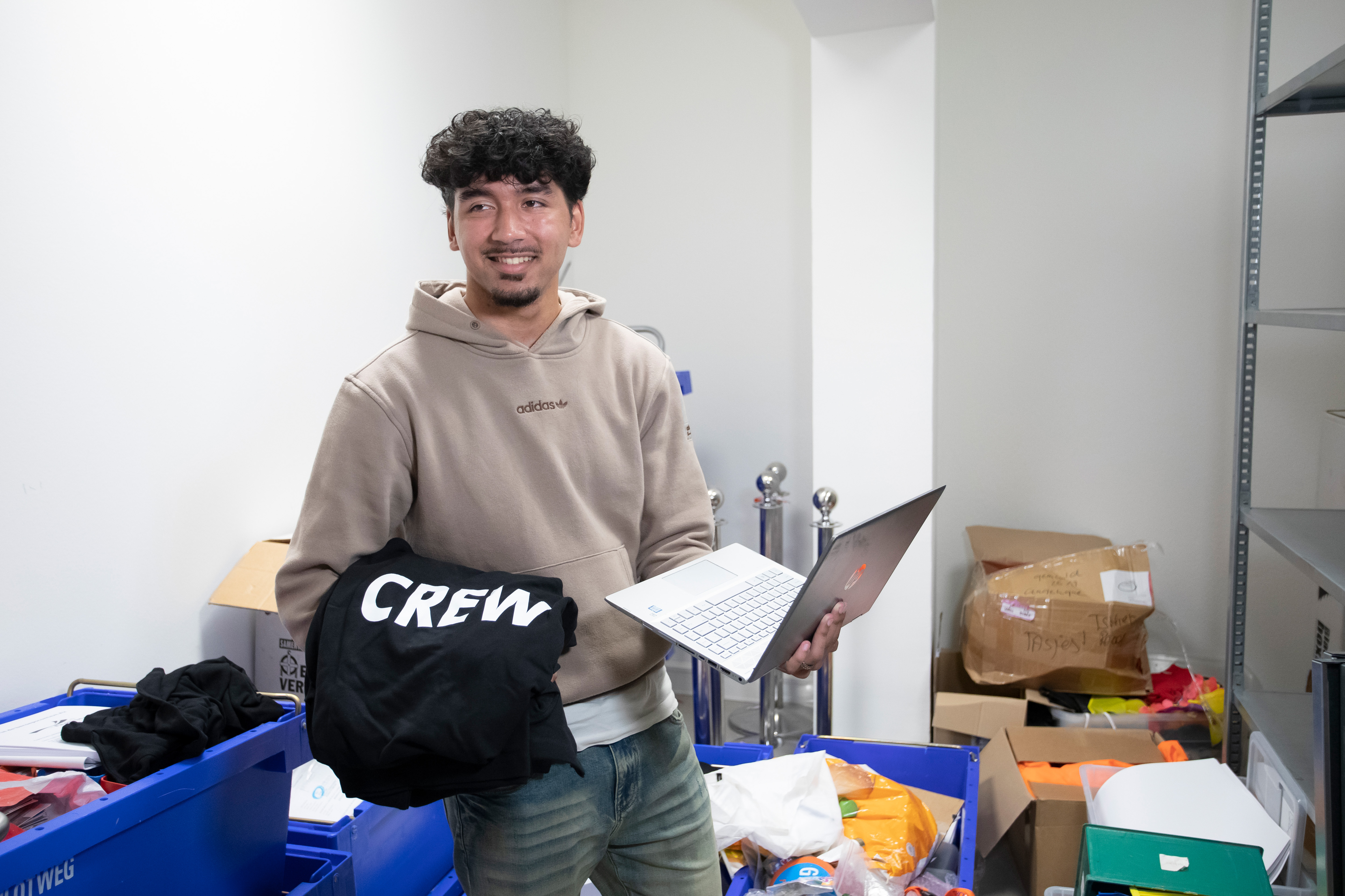 Een jongen staat in een ruimte met allemaal spullen. In zijn ene hand heeft hij een laptop en in zijn andere hand een trui met het woord crew erop