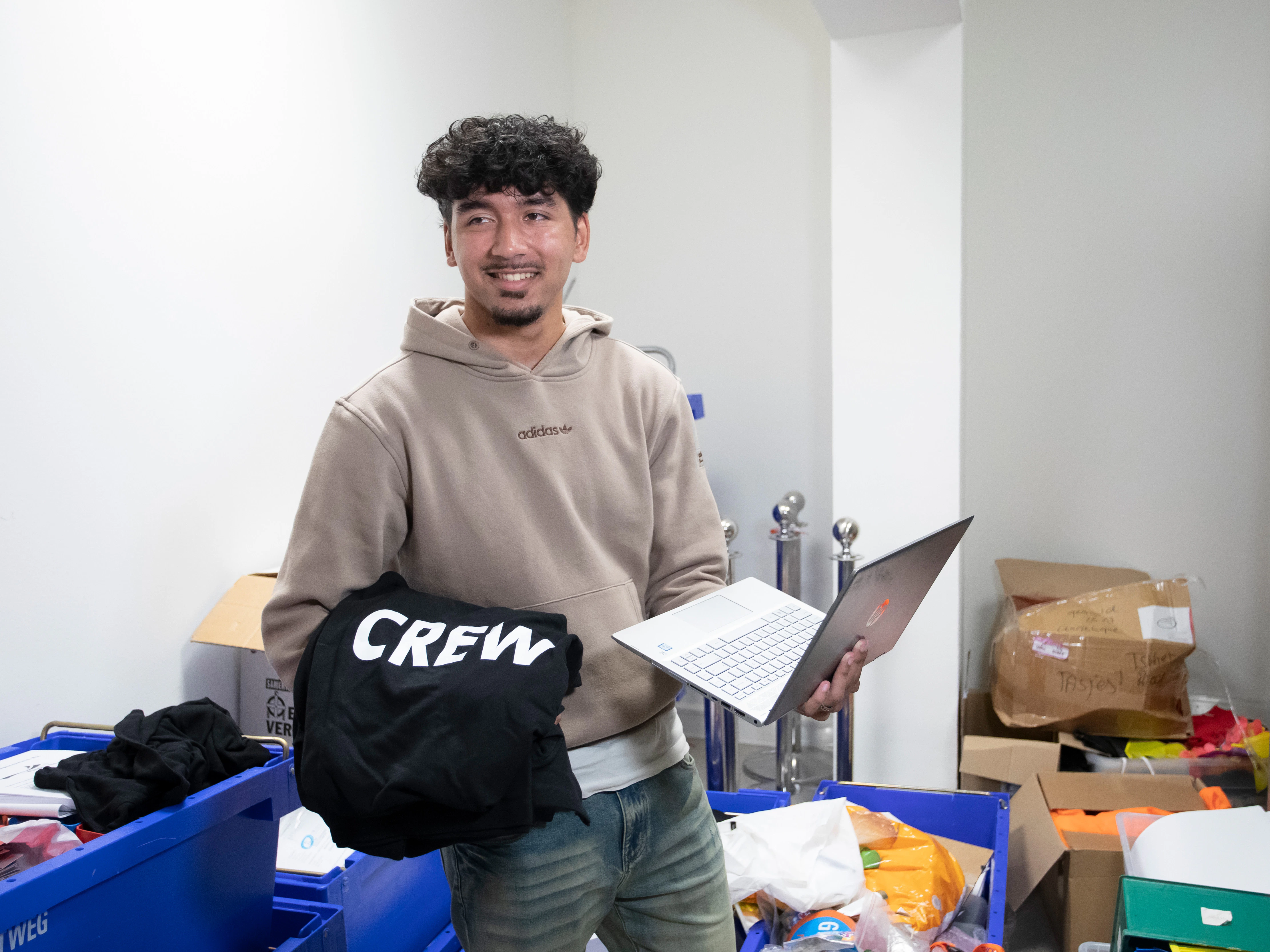 Een jongen staat in een ruimte met allemaal spullen. In zijn ene hand heeft hij een laptop en in zijn andere hand een trui met het woord crew erop