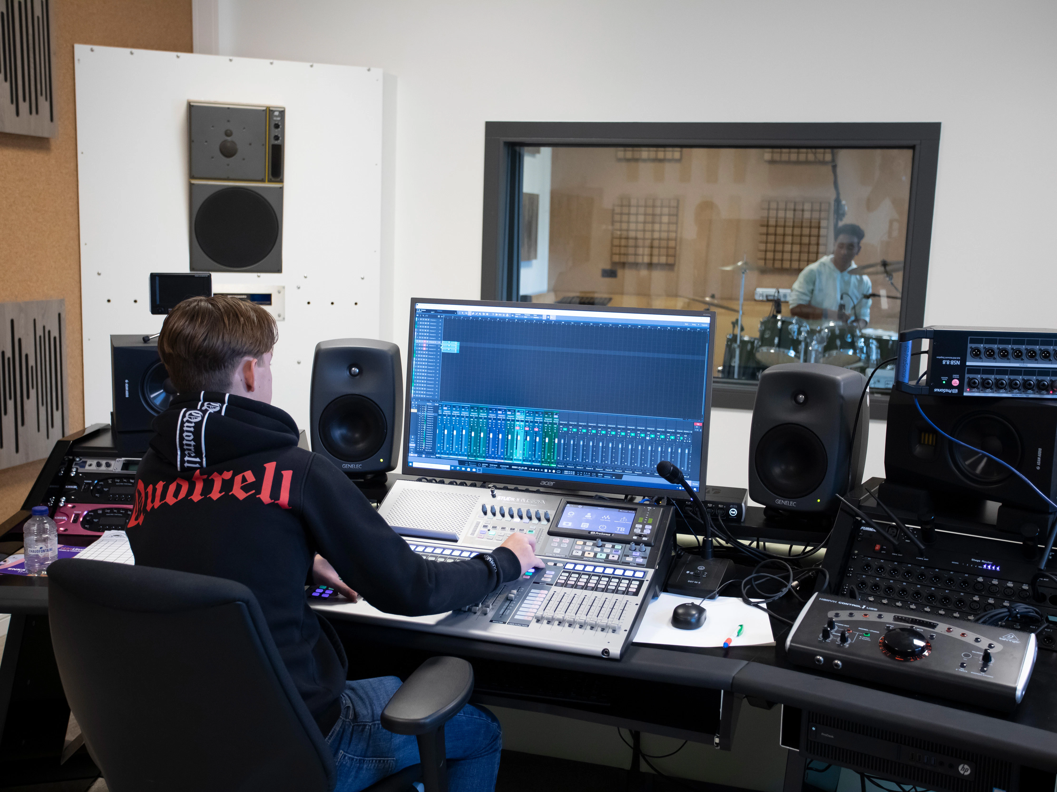 Student zit in een studio achter de computer terwijl een drummer muziek maakt