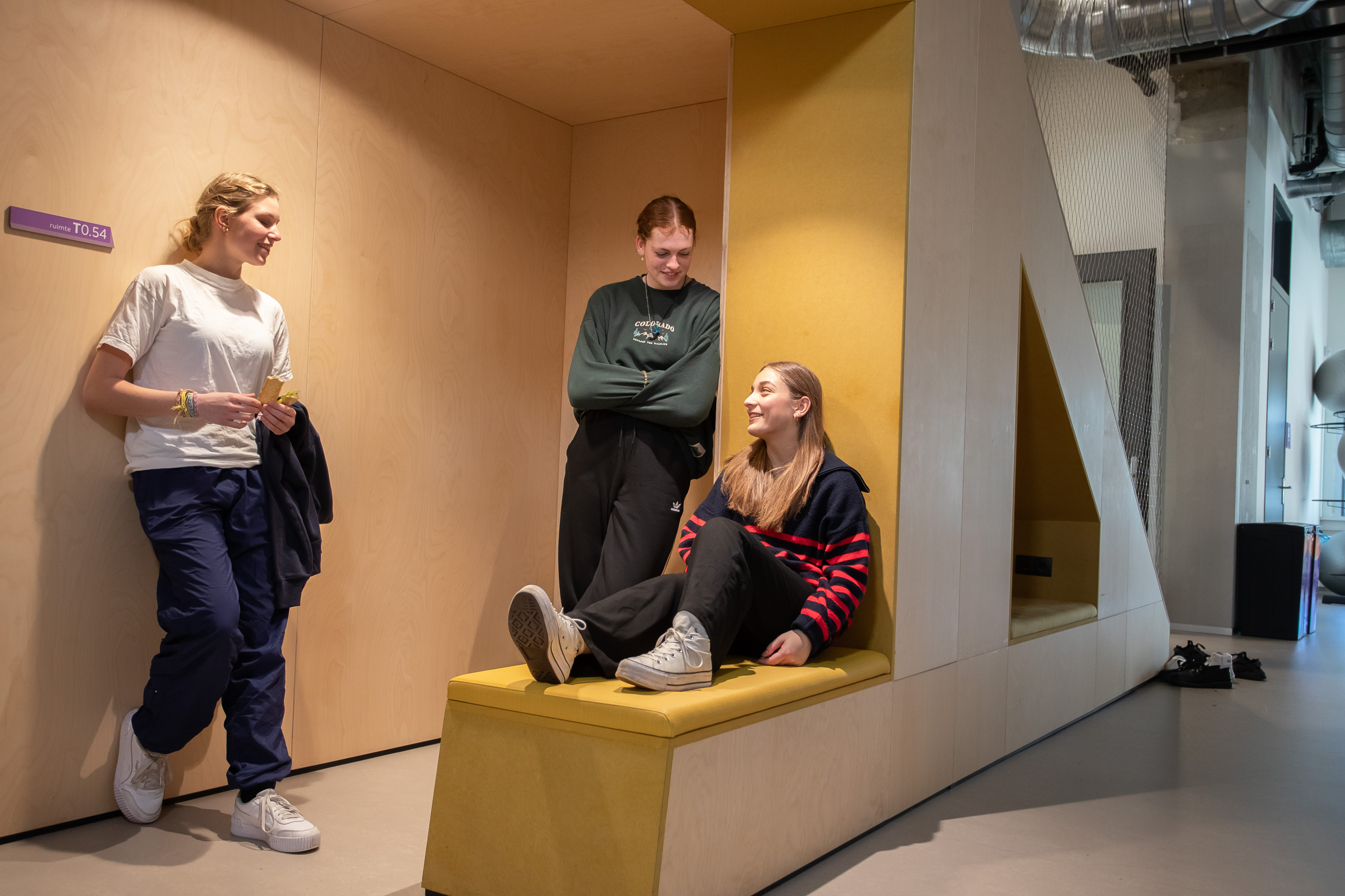 2 studenten staan te wachten voor een lokaal terwijl 1 student op een bankje zit