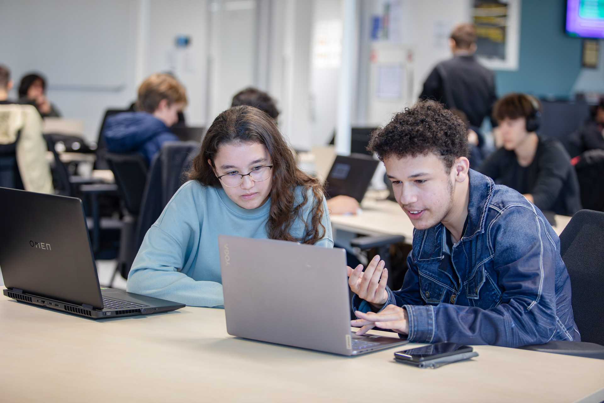 Een jongen en een meisje in een klaslokaal kijken naar een laptop en overleggen met elkaar.