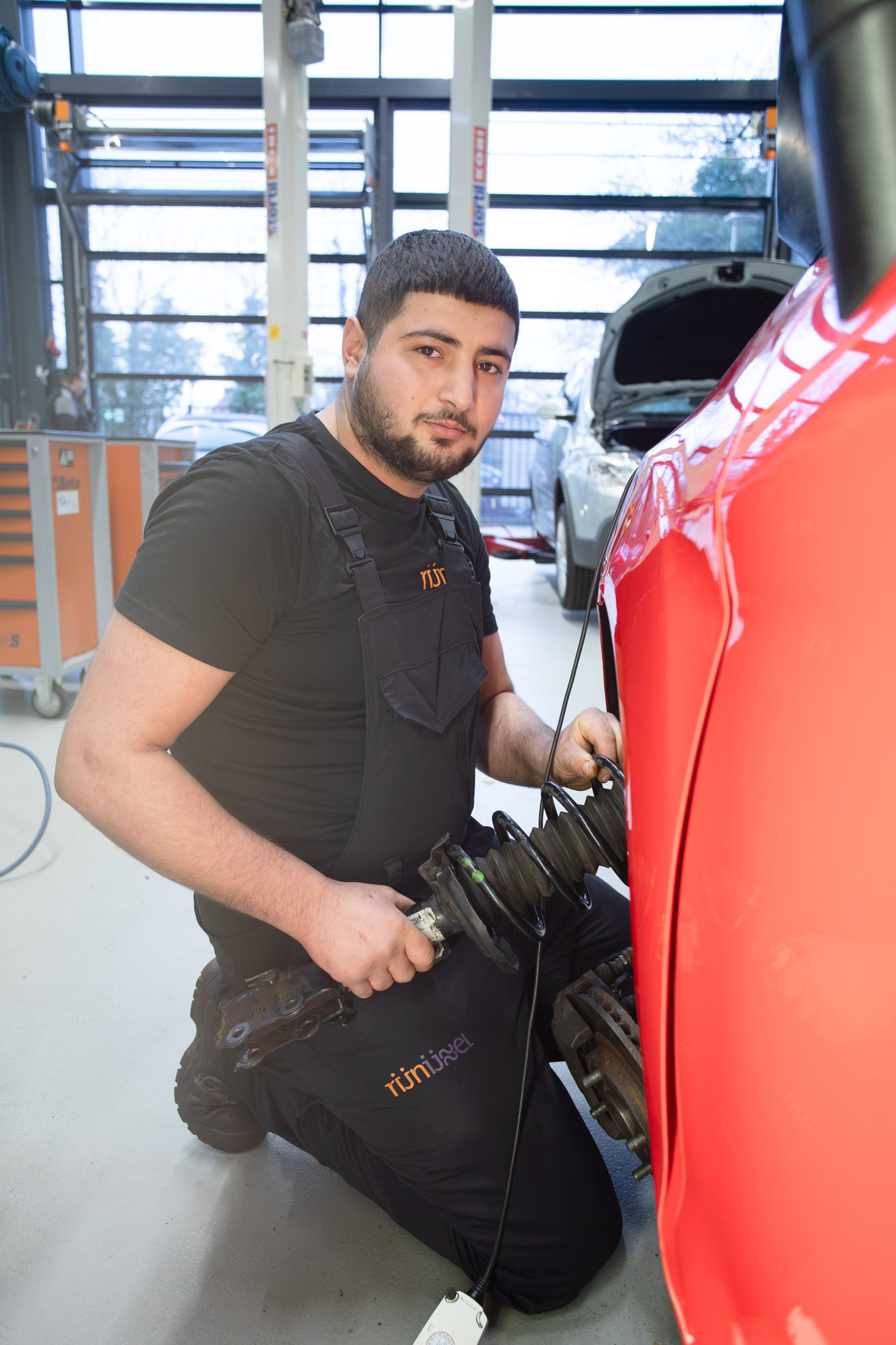 Jongen zit op zijn knieën naast een auto, kijkt in de camera en heeft gereedschap in zijn hand
