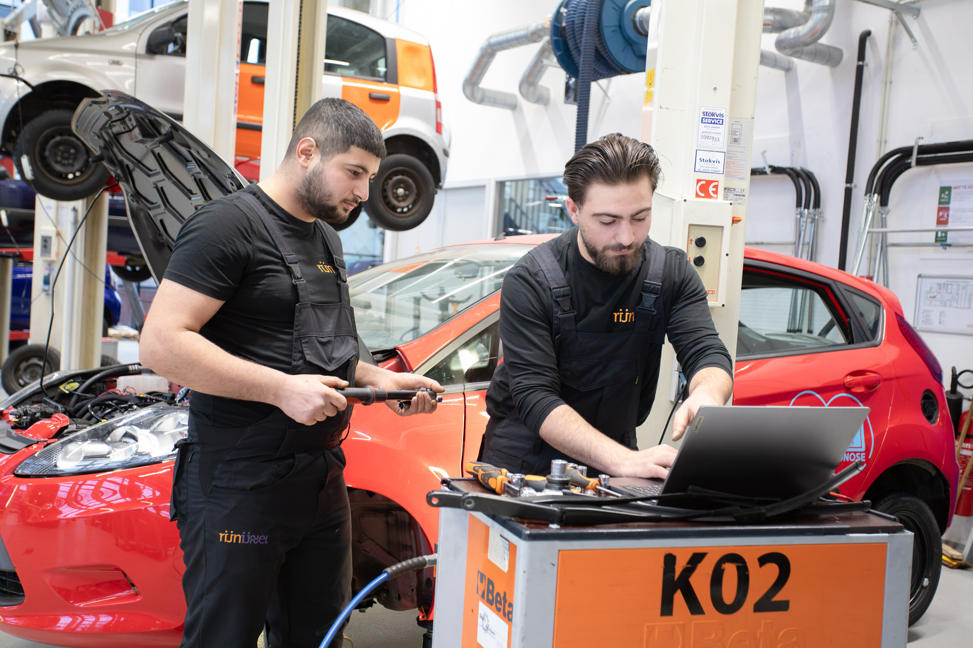 2 mannen inwerkkleding kijken naar een laptop waarvan er een gereedschap in de hand heeft. Op de achtergrond staat een auto met de motorkap open