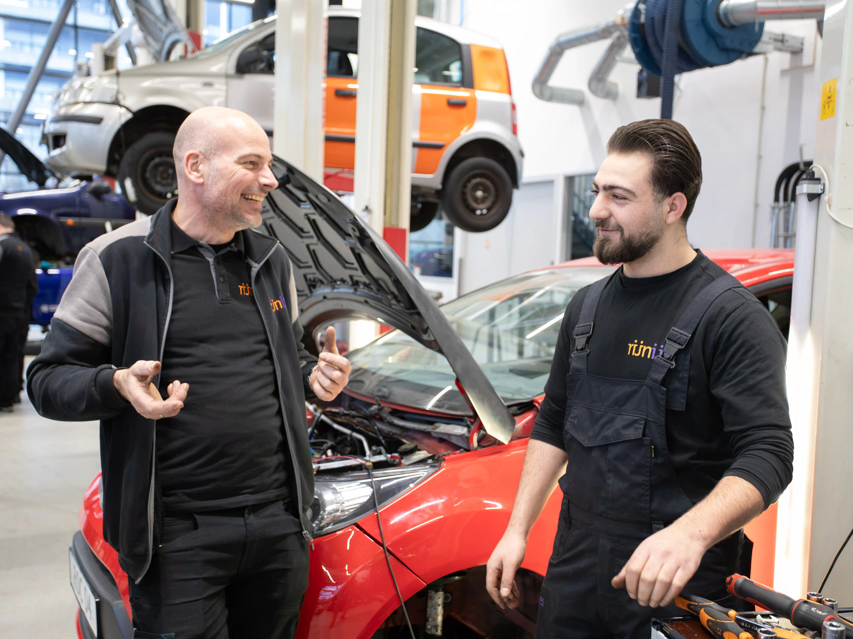2 mannen kijken elkaar lachend aan terwijl ze voor een auto staan met de motorkap open