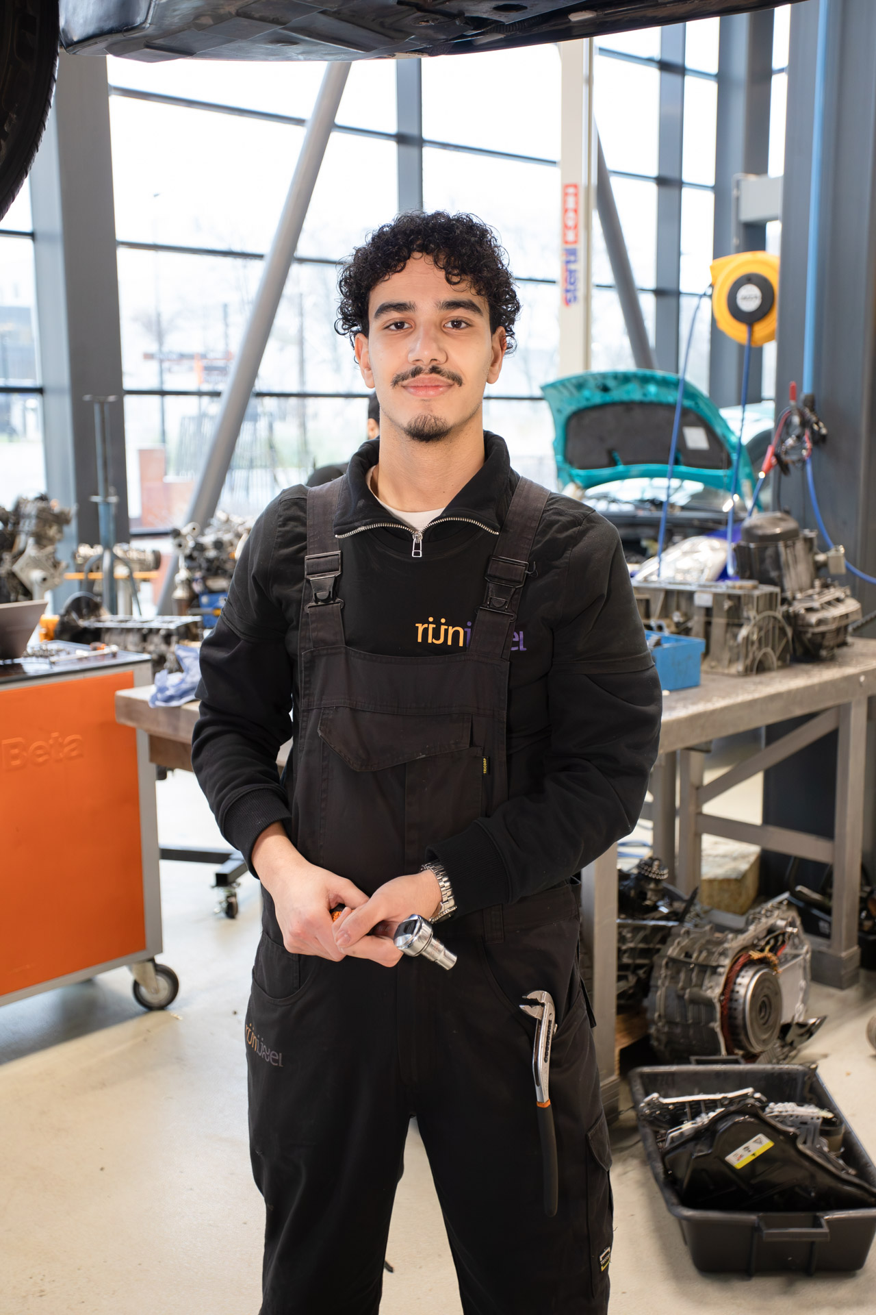 Jongen kijkt in de camera met gereedschap in zijn hand. Op de achtergrond zie je een auto met de motorkap open
