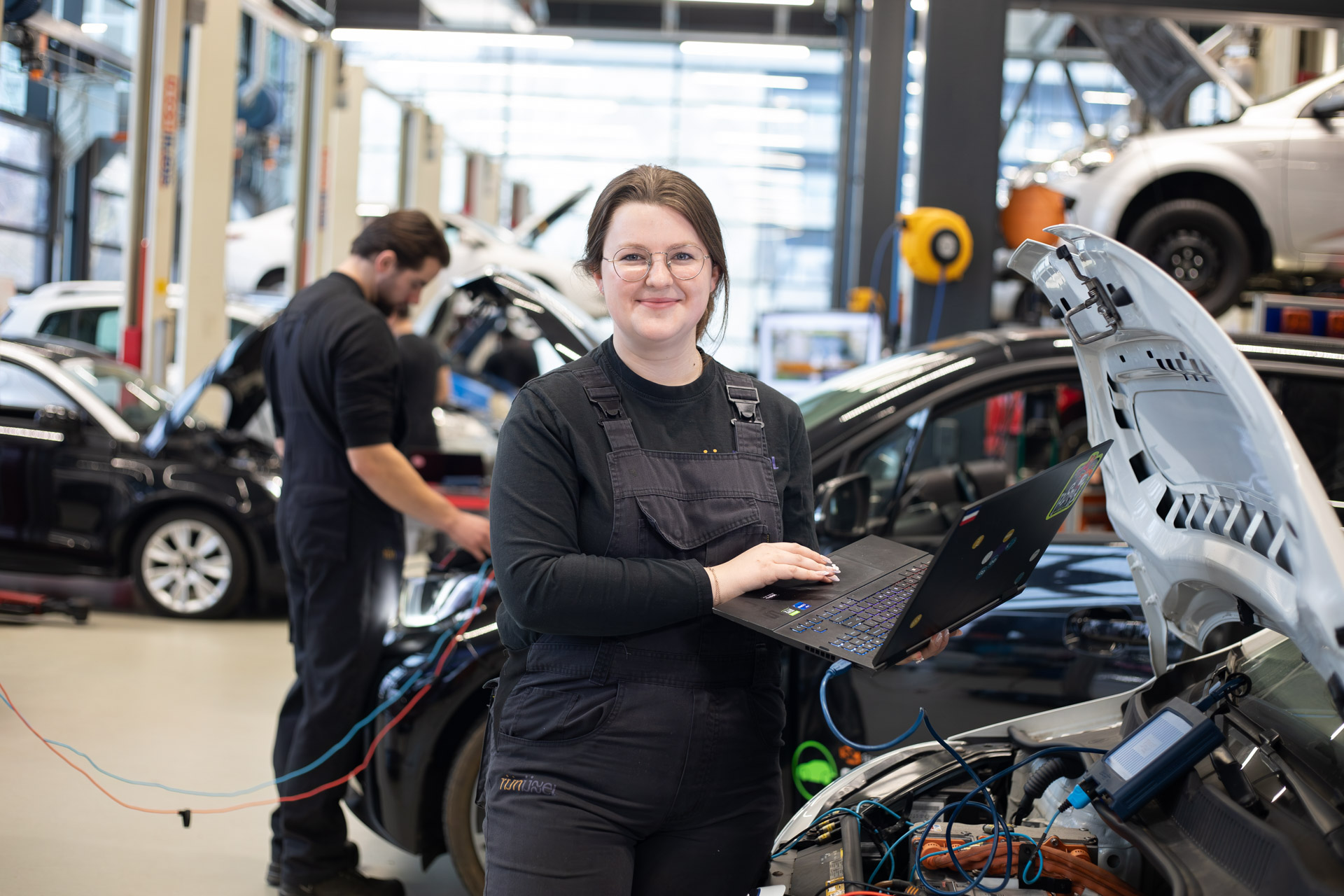 Meisje kijkt in de camera met een laptop in de hand terwijl ze voor een auto staat met de motorkap open