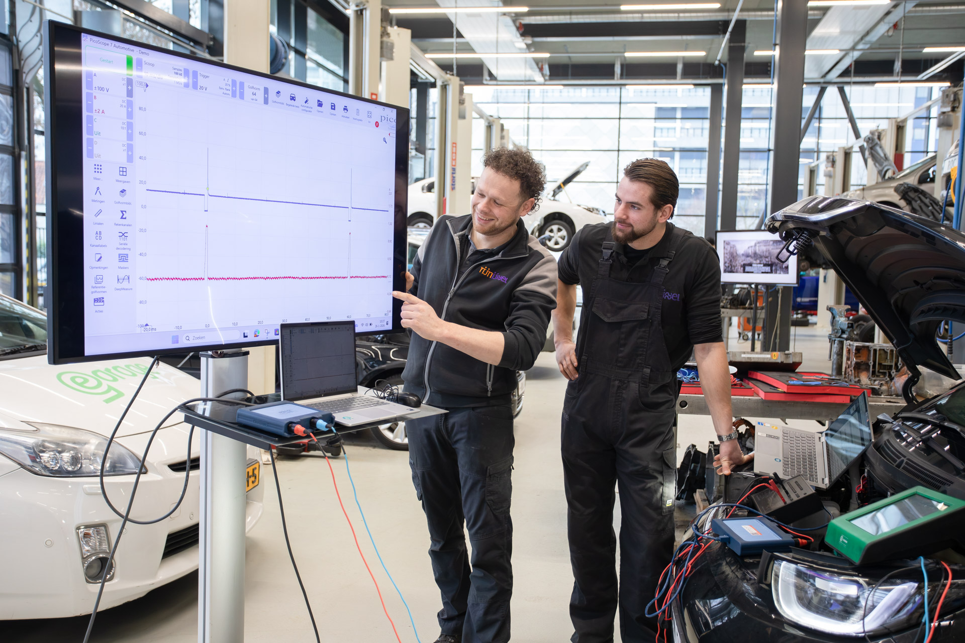 2 mannen kijken naar een televisiescherm waar een overzicht te zien is. Ze staan voor een auto met de motorkap open.