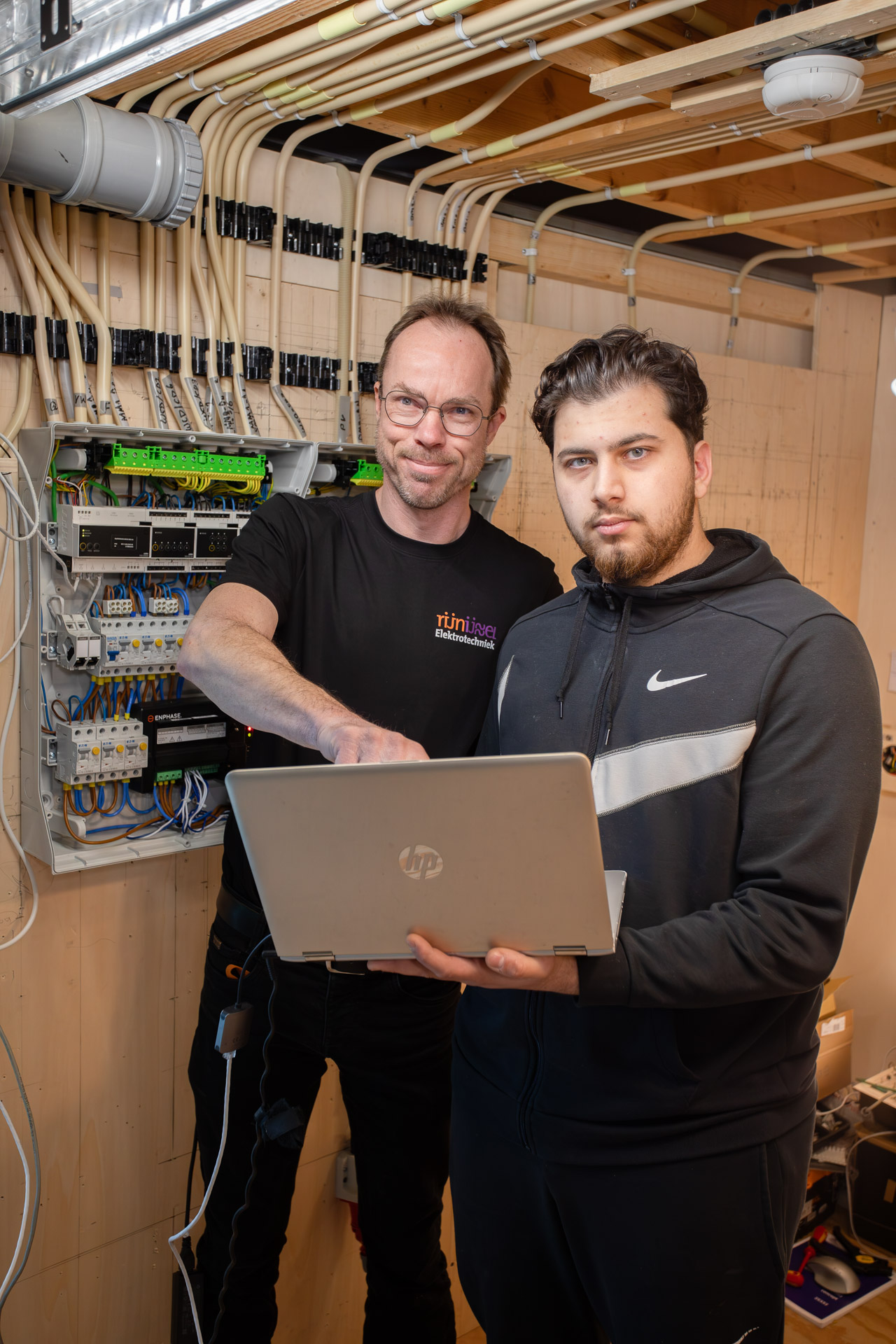 2 mannen kijken in de camera terwijl de een een laptop in zijn hand heeft. Ze staan voor een groepenkast