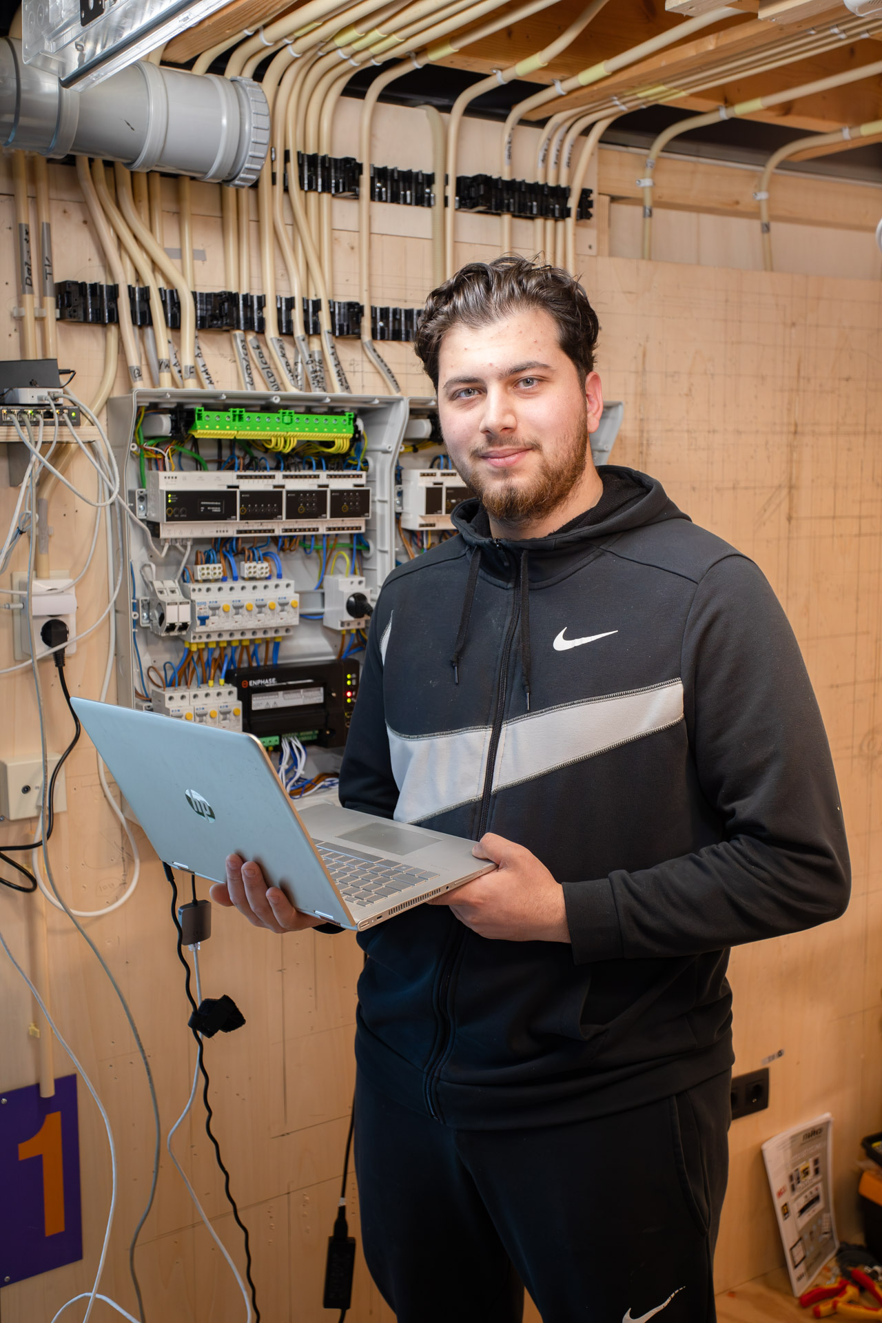 Jongen kijkt in de camera terwijl hij een laptop in zijn hand heeft en voor een groepenkast staat.