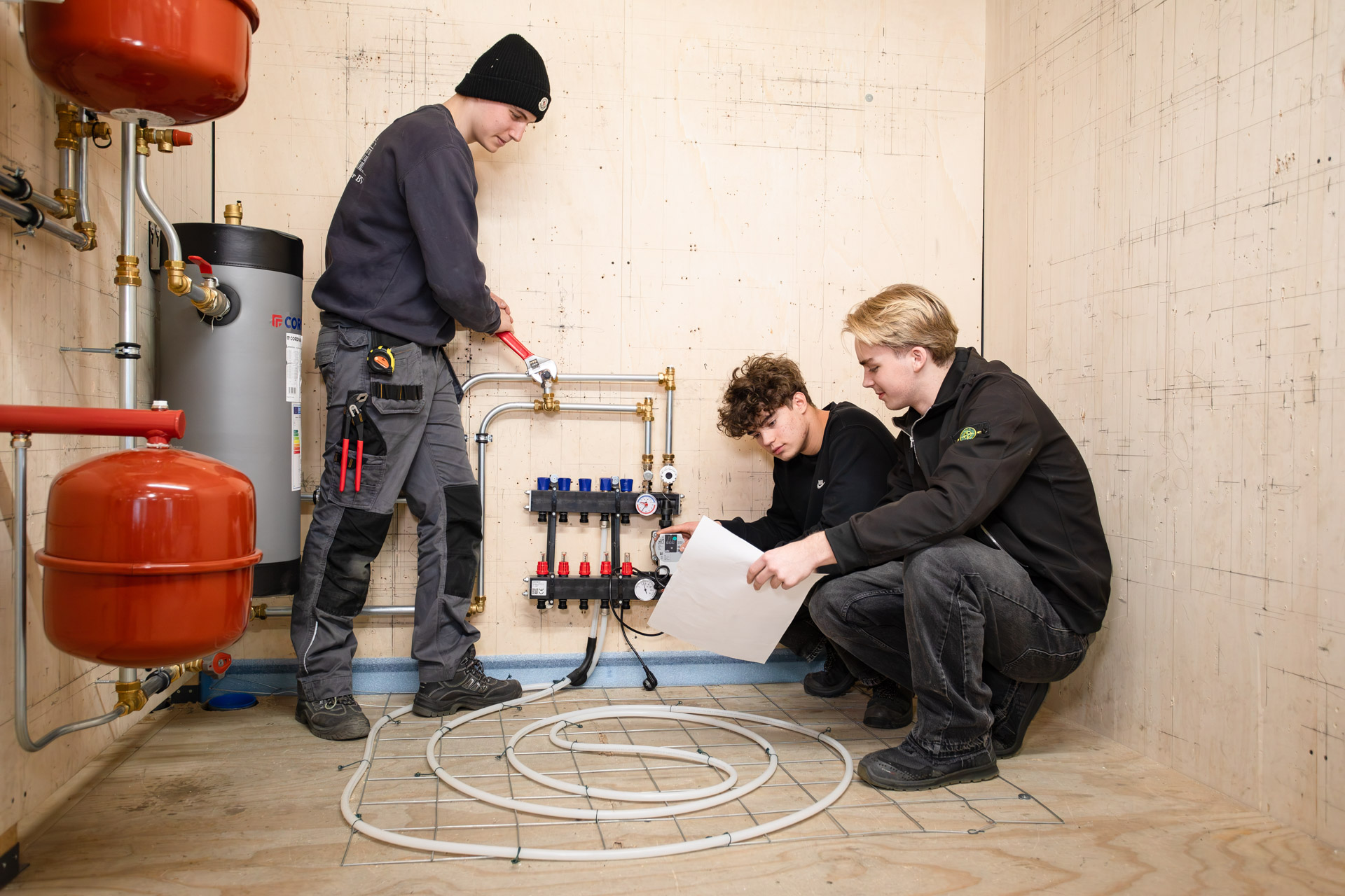 Twee jongens zitten gehurkt op de vloer met vloerverwarming. Een jongen heeft een papiertje in zijn hand en de andere kijkt naar het systeem van de vloerverwarming. Een derde jongen draait met een waterpomptang een koppeling bij de leiding aan.