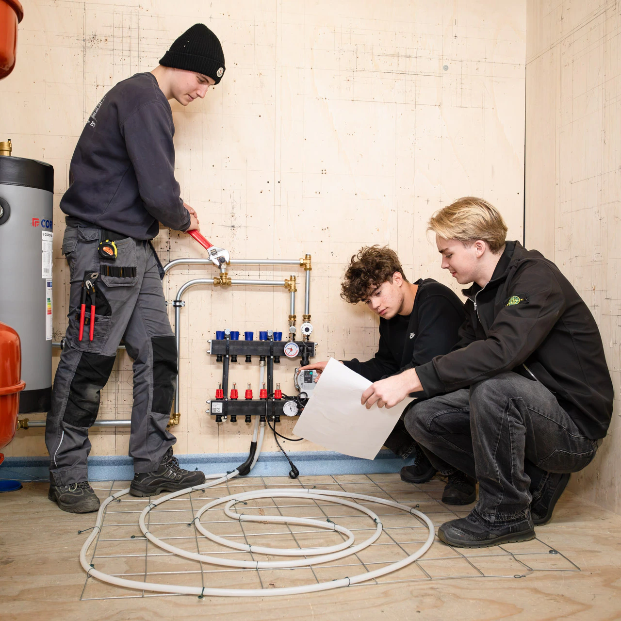 Twee jongens zitten gehurkt op de vloer met vloerverwarming. Een jongen heeft een papiertje in zijn hand en de andere kijkt naar het systeem van de vloerverwarming. Een derde jongen draait met een waterpomptang een koppeling bij de leiding aan.