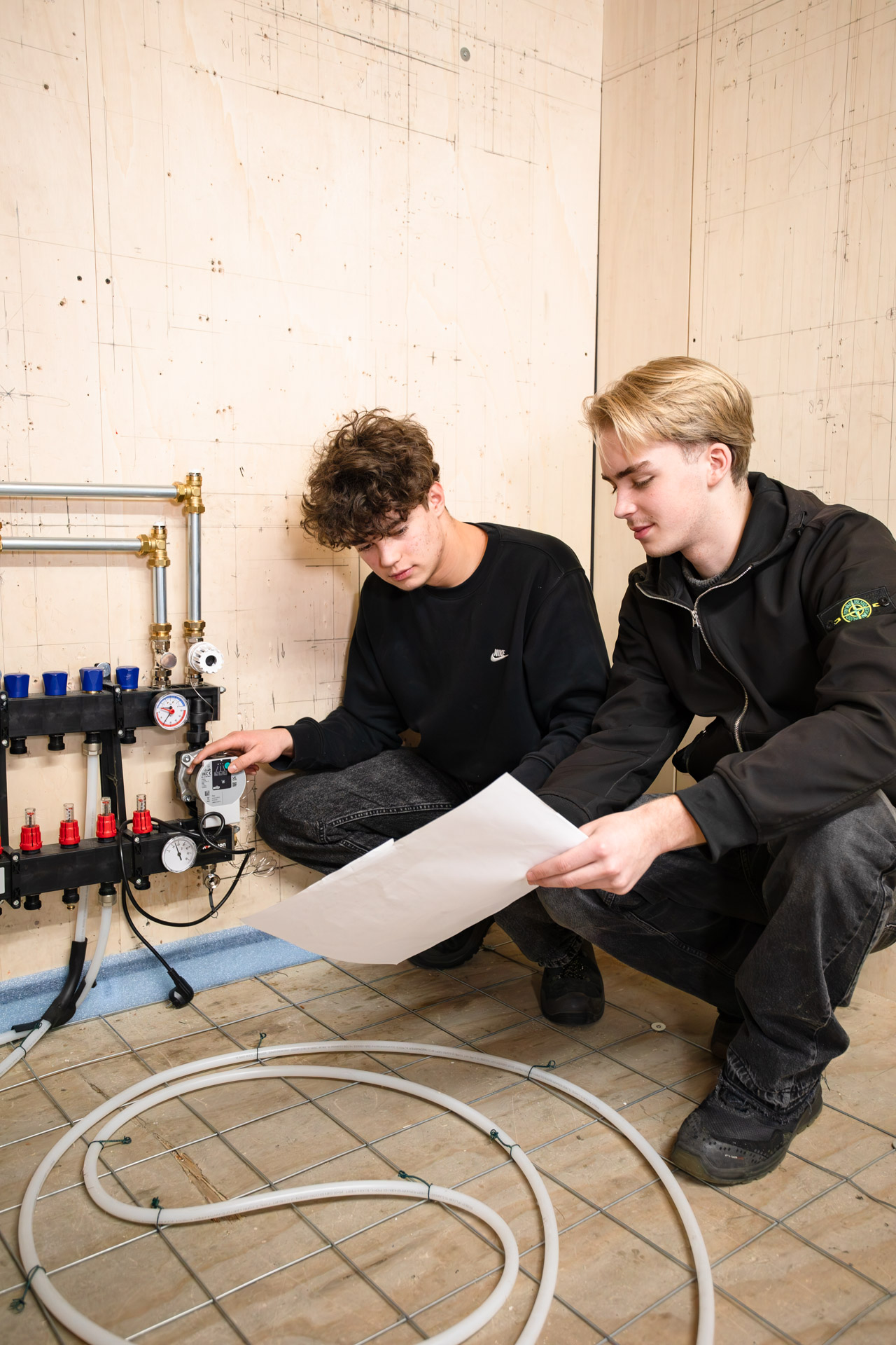 2 jongen zitten gehurkt op een vloer met vloerverwarming. Een jongen heeft een papier in zijn hand en de andere kijkt naar het systeem van de vloerverwarming