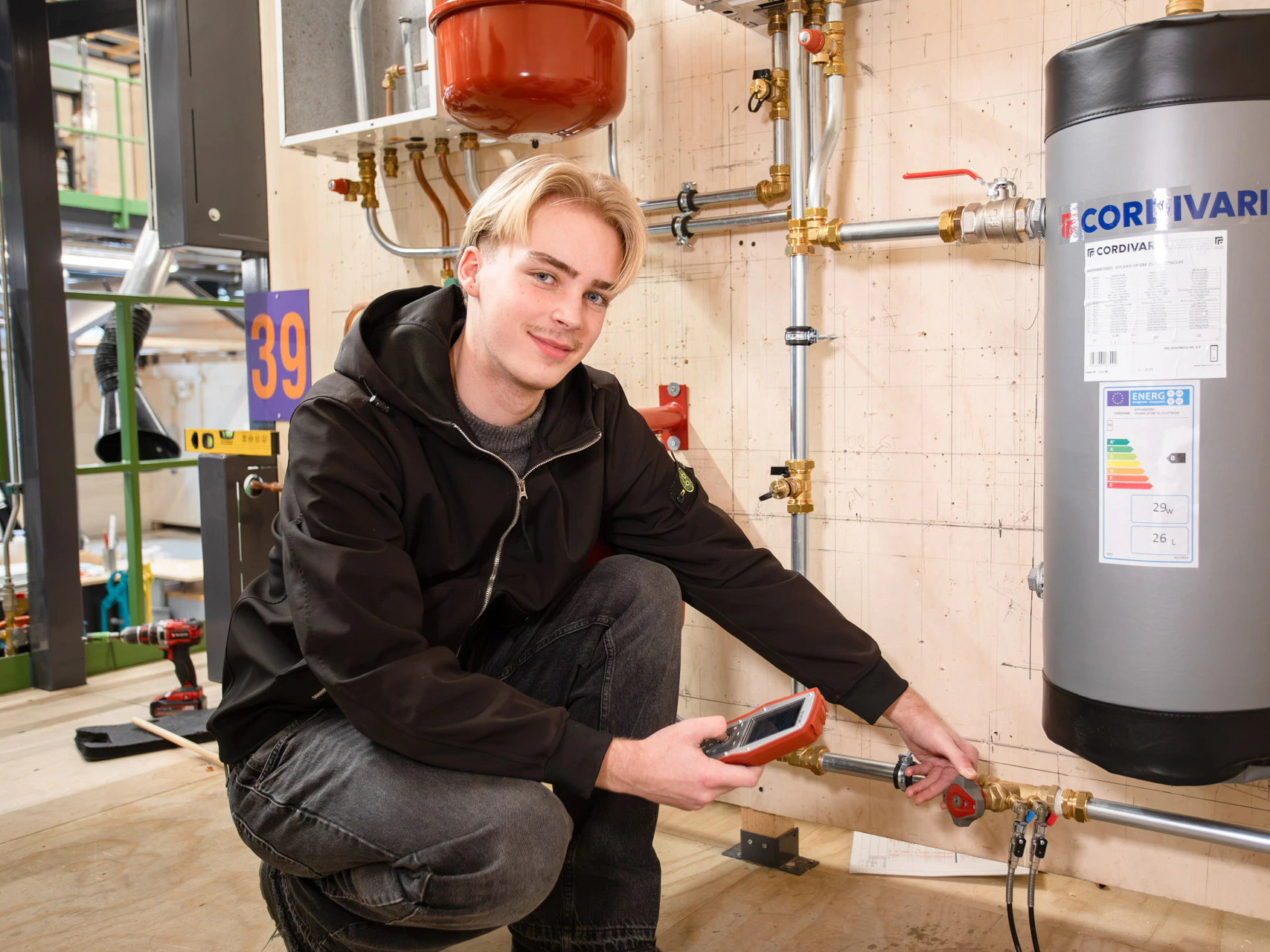 Een jongen kijkt in de camera terwijl hij voor een cv ketel zit met meetapparatuur in zijn hand