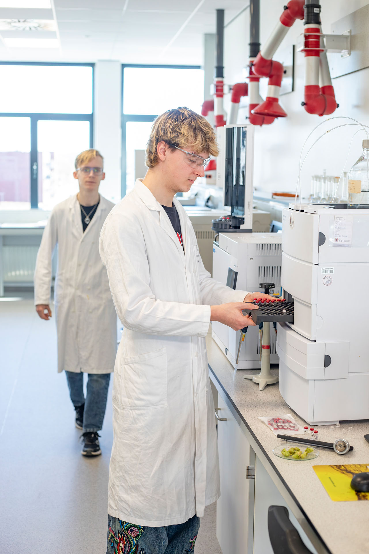 Een jongen is bezig met buisjes uit een apparaat halen in een laboratorium terwijl op de achtergrond een jongen komt aanlopen